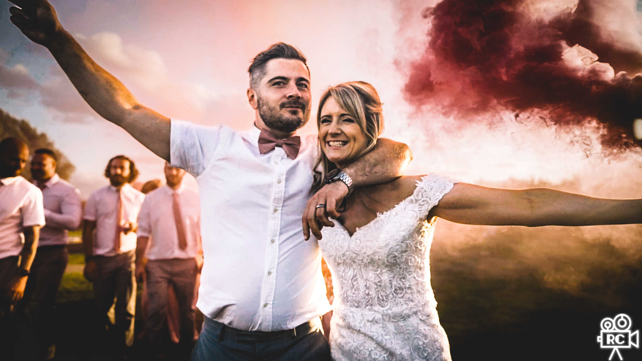 Joyful wedding couple embracing with pink smoke, groomsmen behind, warm sunset light and lively outdoor reception atmosphere.