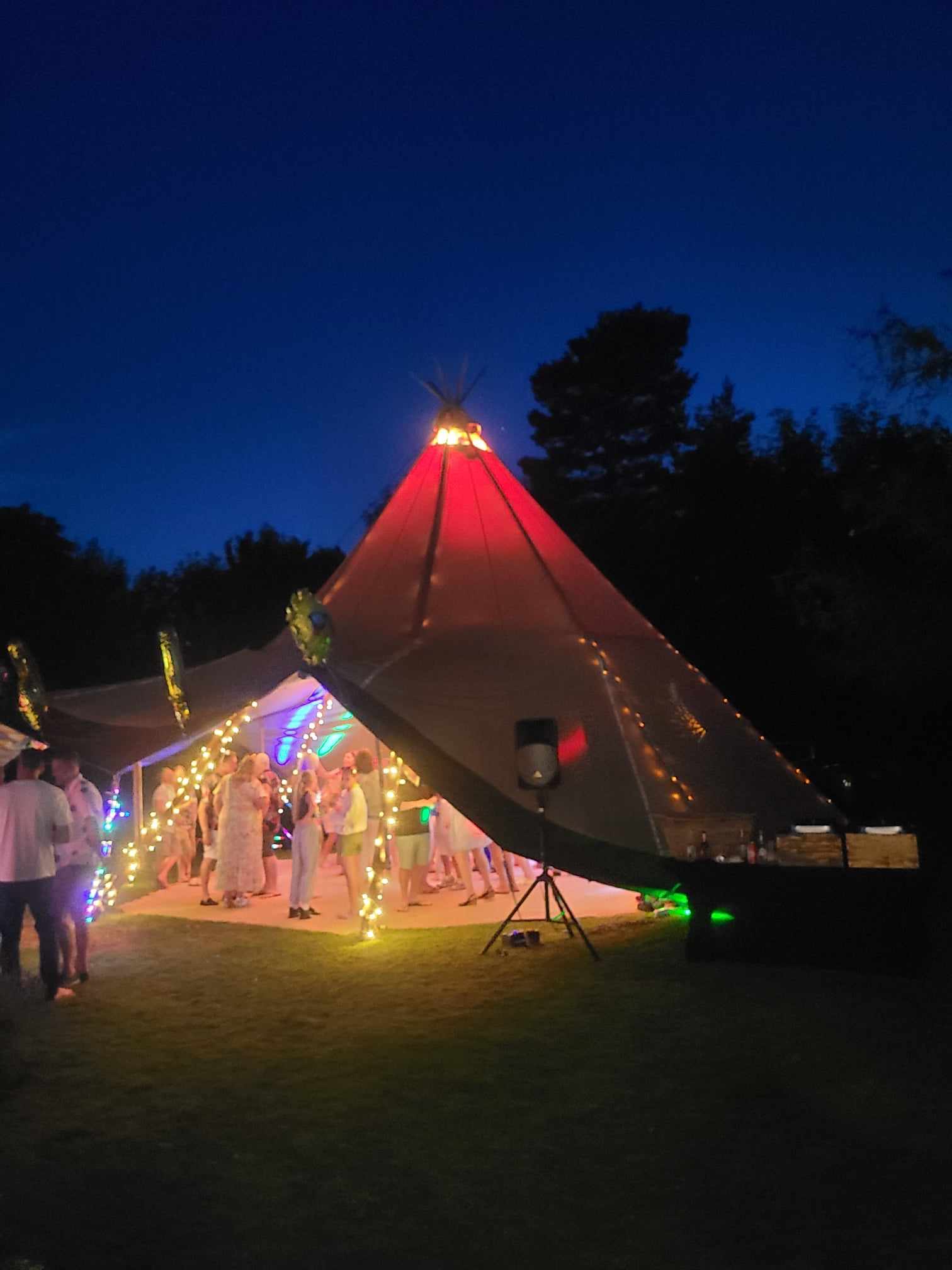 A vibrant tent glows with multicolored lights, hosting guests in white attire dancing under starry skies, exuding a festive atmosphere.