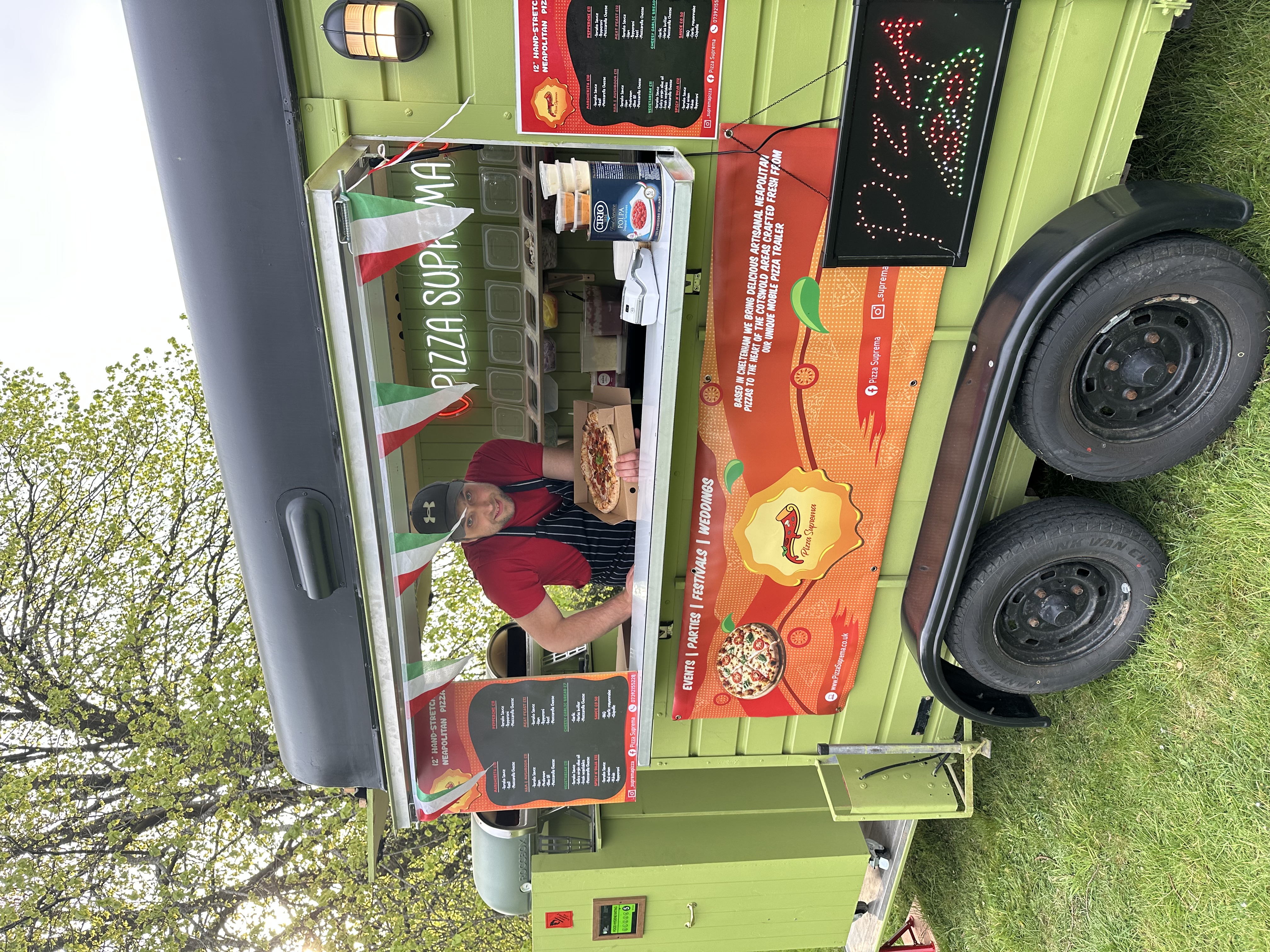 A friendly chef in a striped apron showcases a freshly baked pizza from a vintage green food truck adorned with colorful bunting and menu boards.