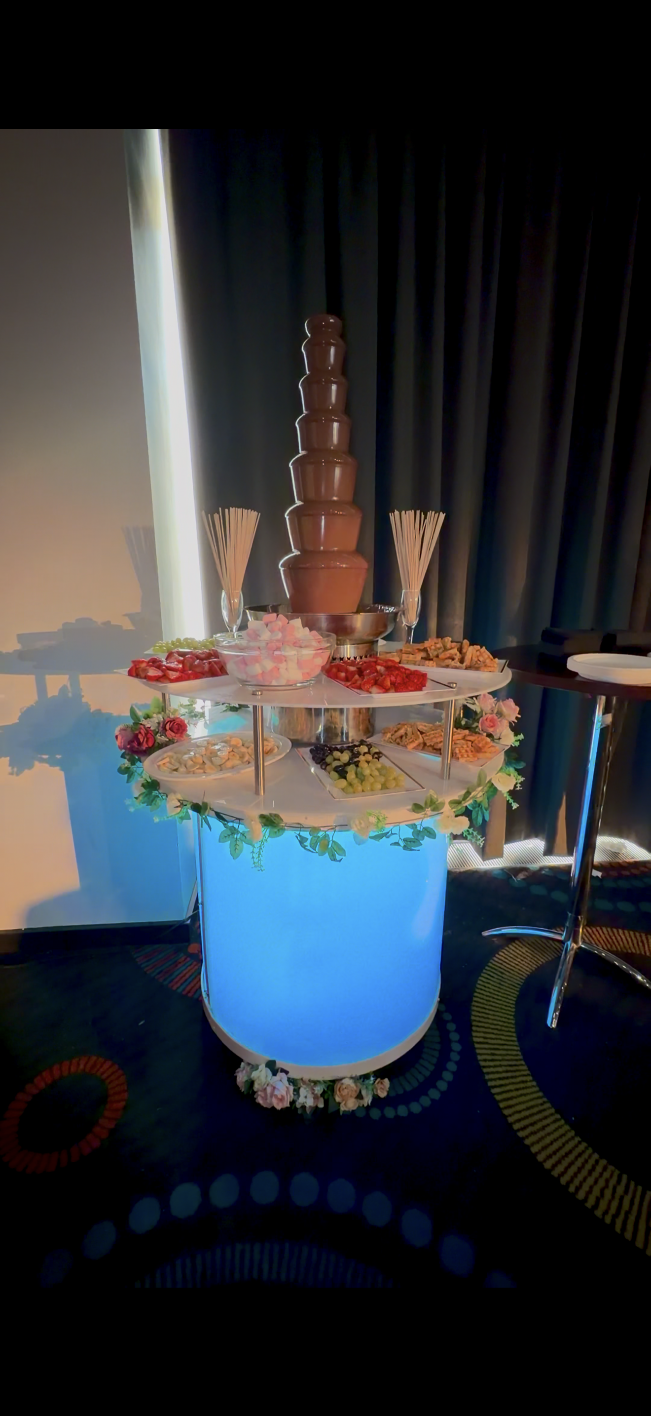 Elegant catering chocolate fountain atop glowing blue buffet, ringed with strawberries, grapes, marshmallows and wafers, warm event lighting.