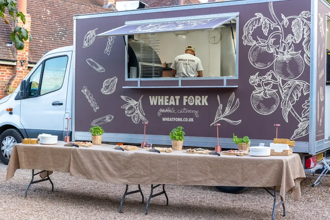 Wheat Fork catering van with chalkboard-style graphics, serving a linen-covered buffet of pastries, herbs and plates in bright daylight.