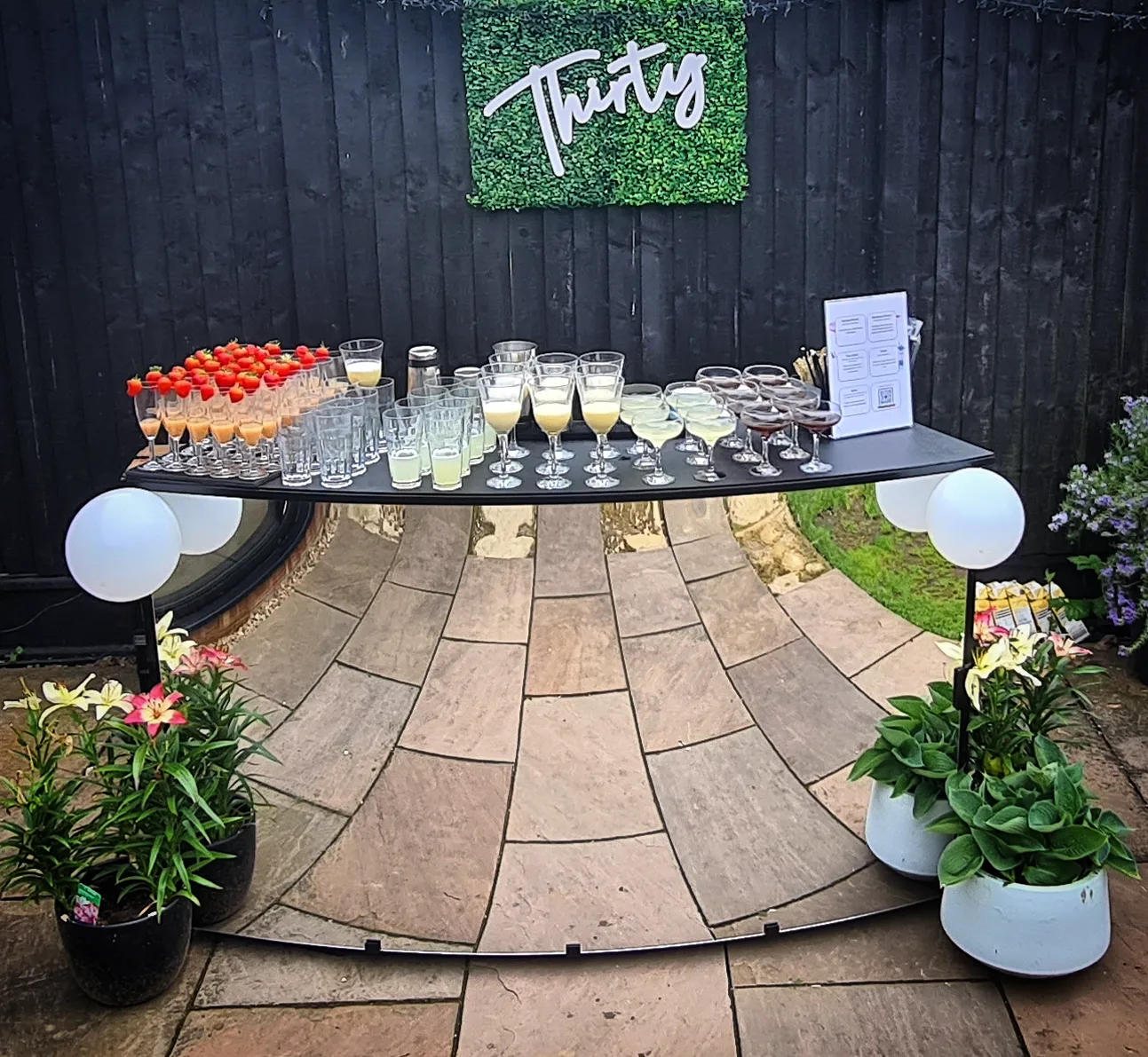 A stylish black table showcases an array of colorful cocktails in elegant glassware, surrounded by vibrant flowers, under bright, inviting daylight.