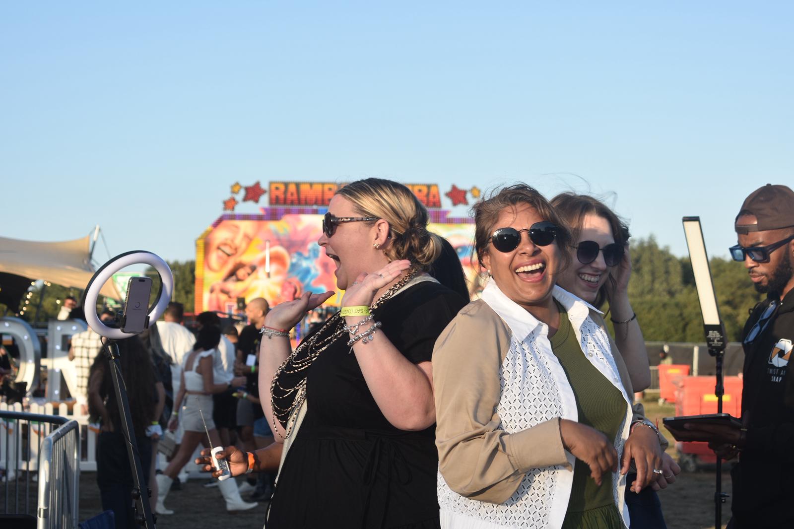 In a vibrant outdoor festival, joyful women in stylish outfits laugh and pose under bright daylight, surrounded by colorful signage and a lively crowd.