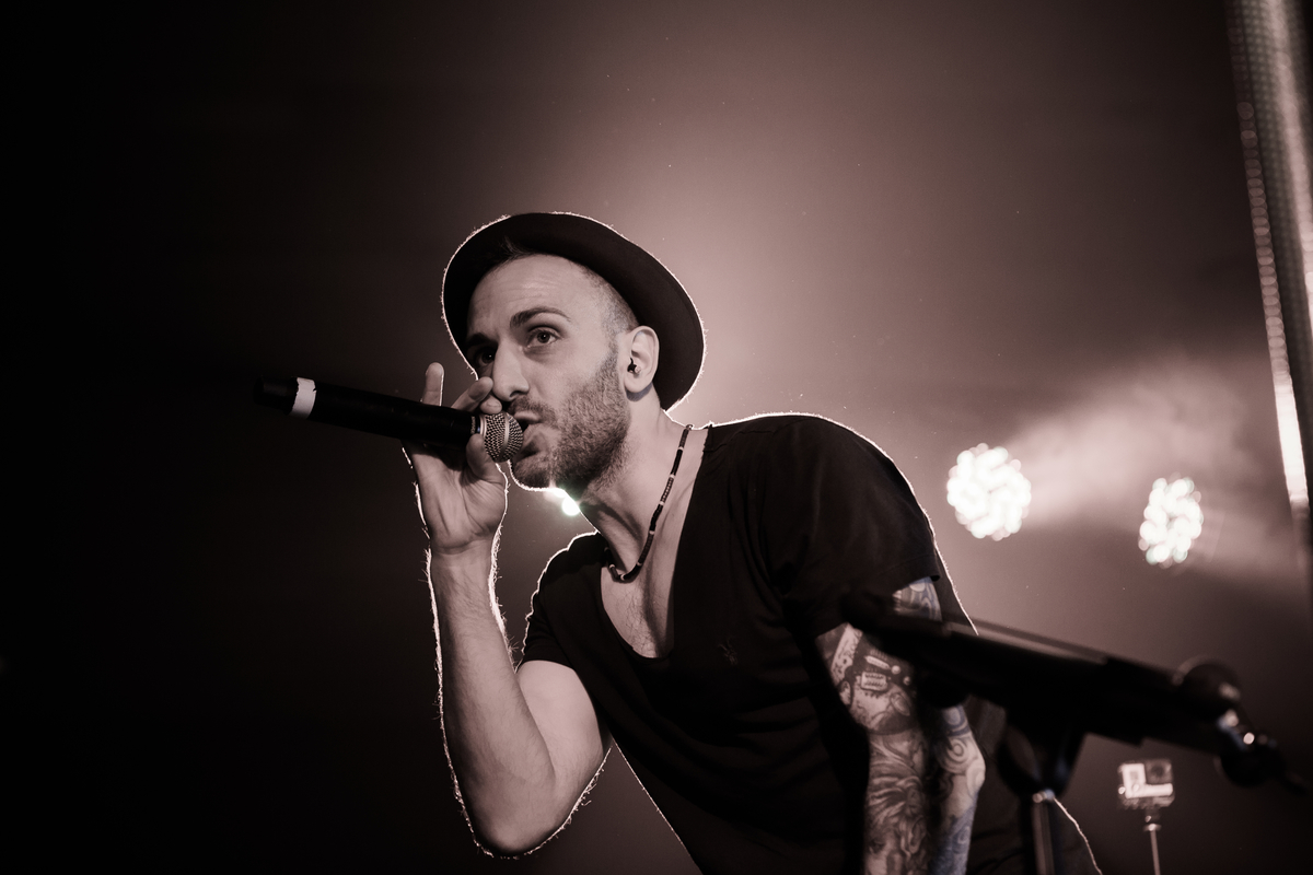 A vibrant performer in a black shirt and hat, passionately singing into a microphone under dynamic stage lighting, creating an energetic atmosphere.