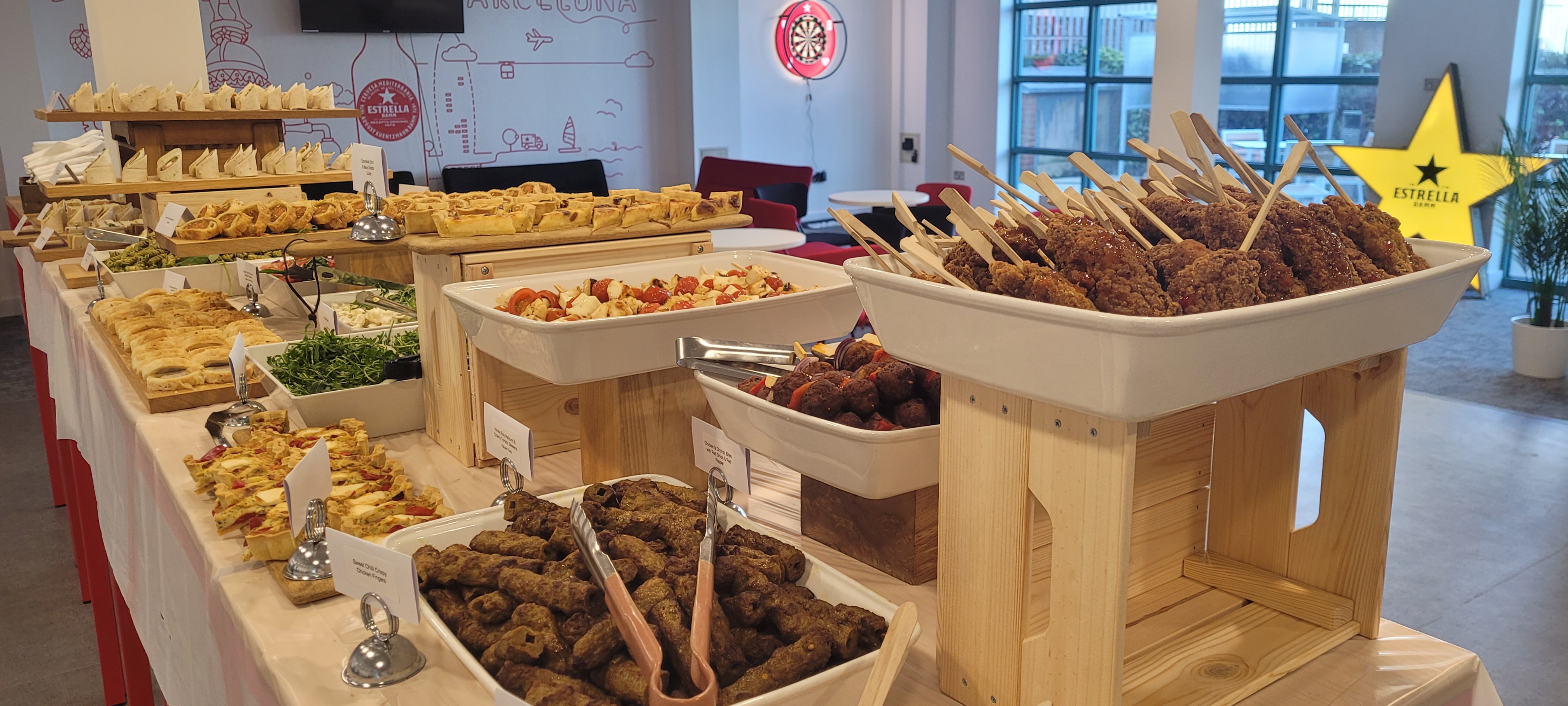 A vibrant catering buffet displays an array of dishes—crispy skewered meats, fresh salads, and pastries—on rustic wooden stands, illuminated by bright daylight.