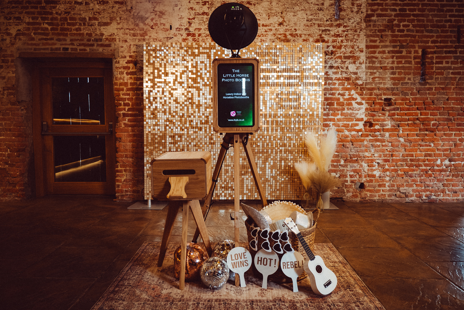 The stylish photo booth features a mosaic backdrop, wooden tripod, and decorative props like disco balls and playful signs, creating a lively atmosphere.