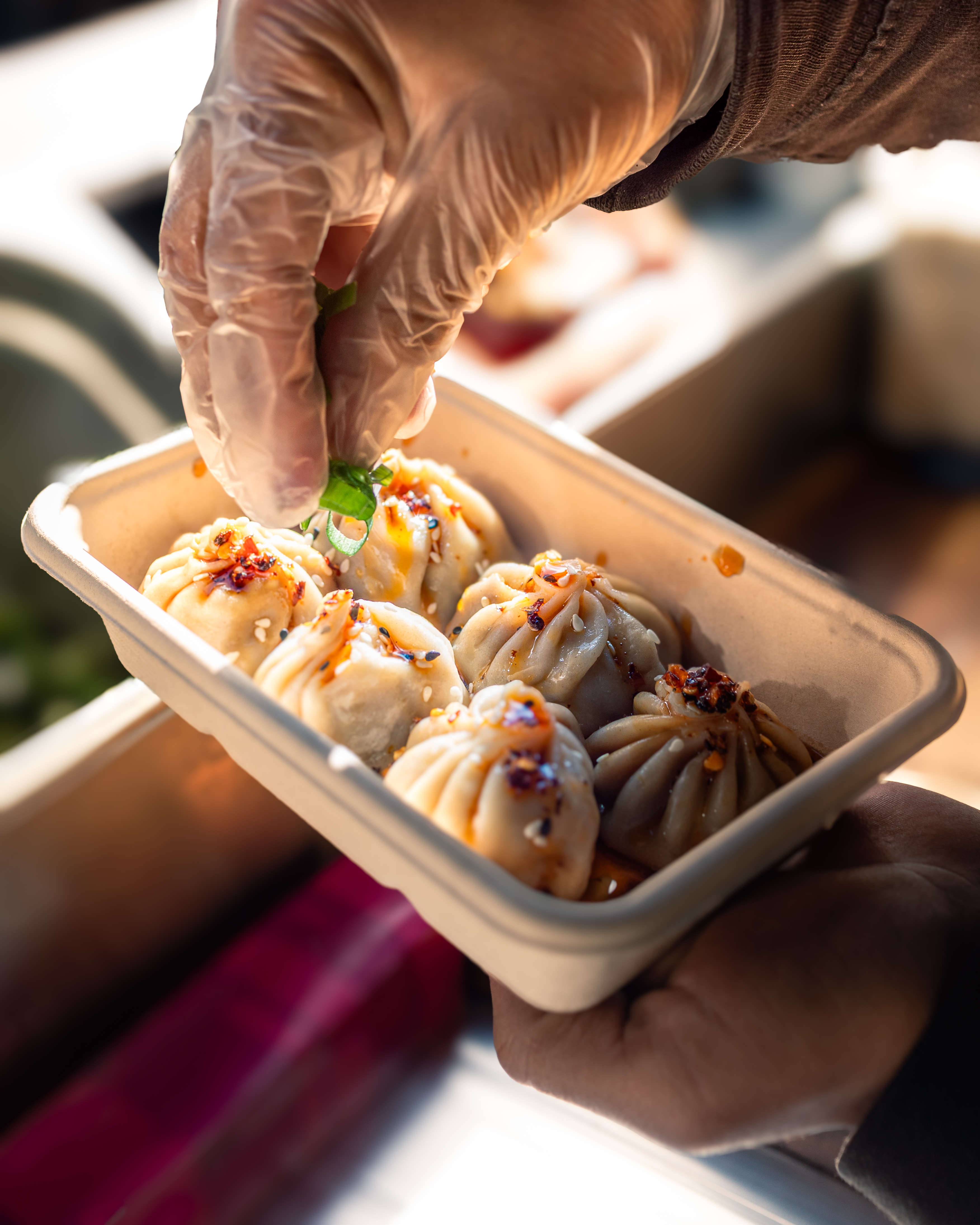 A hand in a glove garnishes steamed dumplings in a natural kraft box, showcasing vibrant colors and an inviting, fresh presentation against a bright, sunny backdrop.