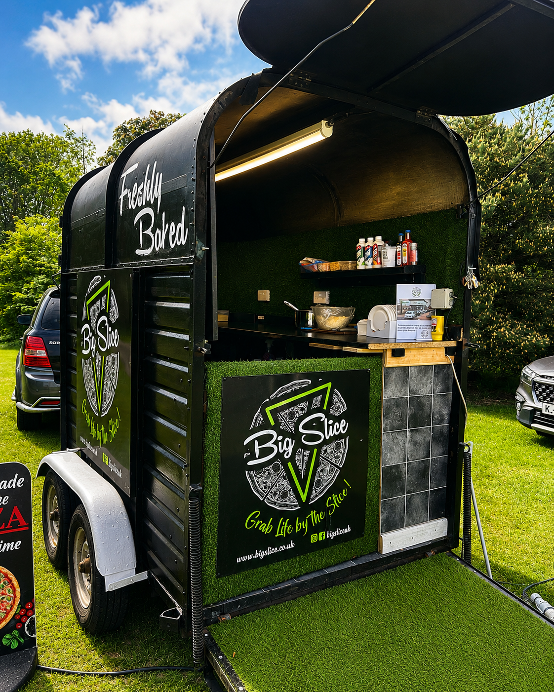 The vibrant black food trailer features a grassy entrance and chalkboard menus, set against a rustic brick wall, inviting hungry patrons.