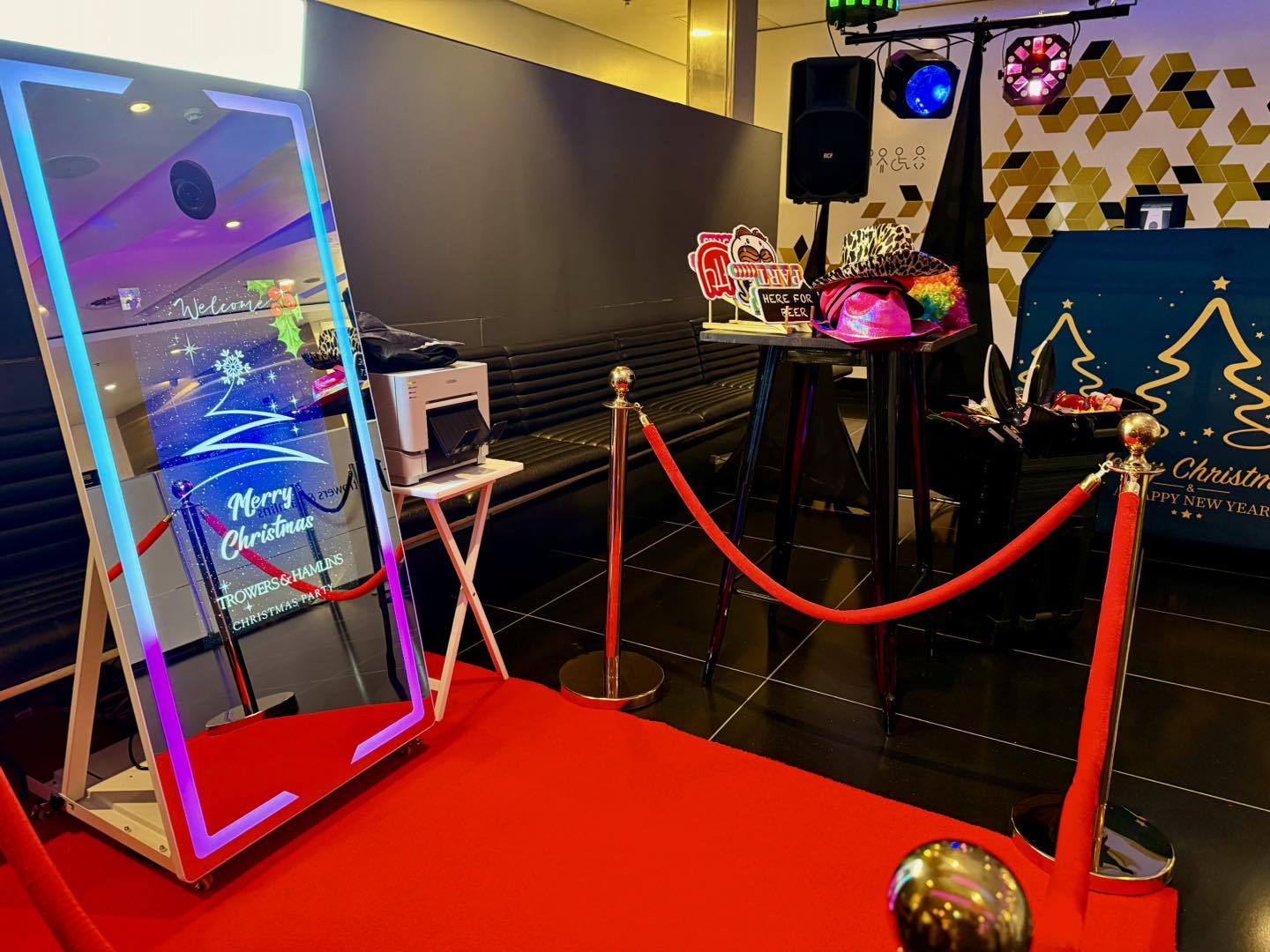 A red carpet leads to a glowing mirror framed in neon, surrounded by polished gold stanchions; twinkling string lights create a festive, glamorous atmosphere perfect for events.