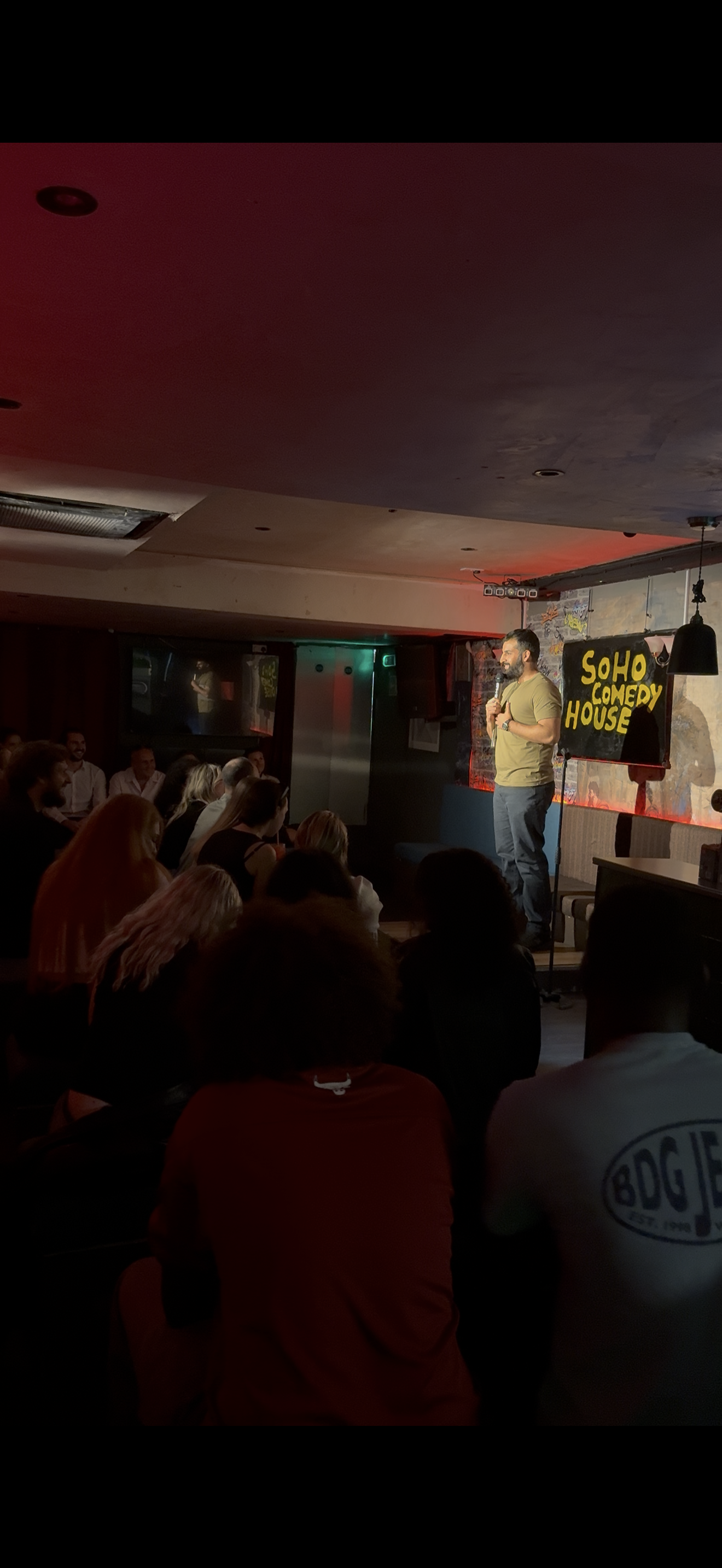 Crowded Soho Comedy House, a stand-up comic with mic on a small stage, red-and-amber lighting, intimate club vibes.