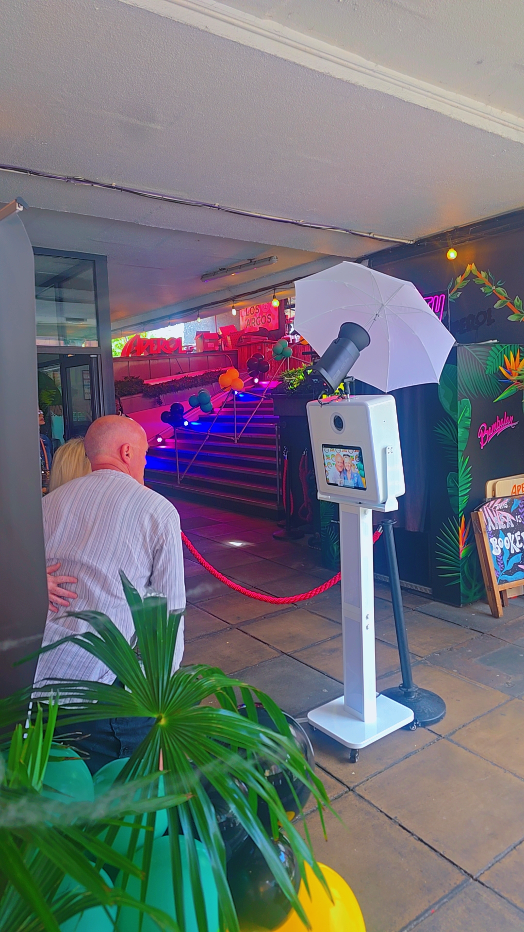 Colorful event photo booth with white umbrella flash, tablet kiosk, red rope stanchions, and neon-lit stairs at a lively catering venue.