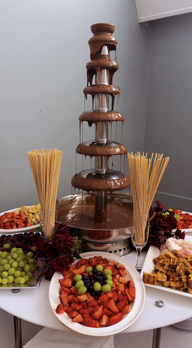 A sleek chocolate fountain stands atop a glowing cylindrical base, surrounded by fresh strawberries, grapes, and marshmallows, creating an inviting dessert display.