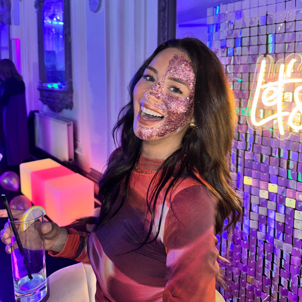 Smiling woman with pink glitter face paint holds a cocktail by a neon mosaic wall, glowing purple-pink nightclub catering vibe.