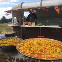 A vibrant outdoor buffet features two large paella pans brimming with saffron rice, bell peppers, and peas, glowing under bright daylight.
