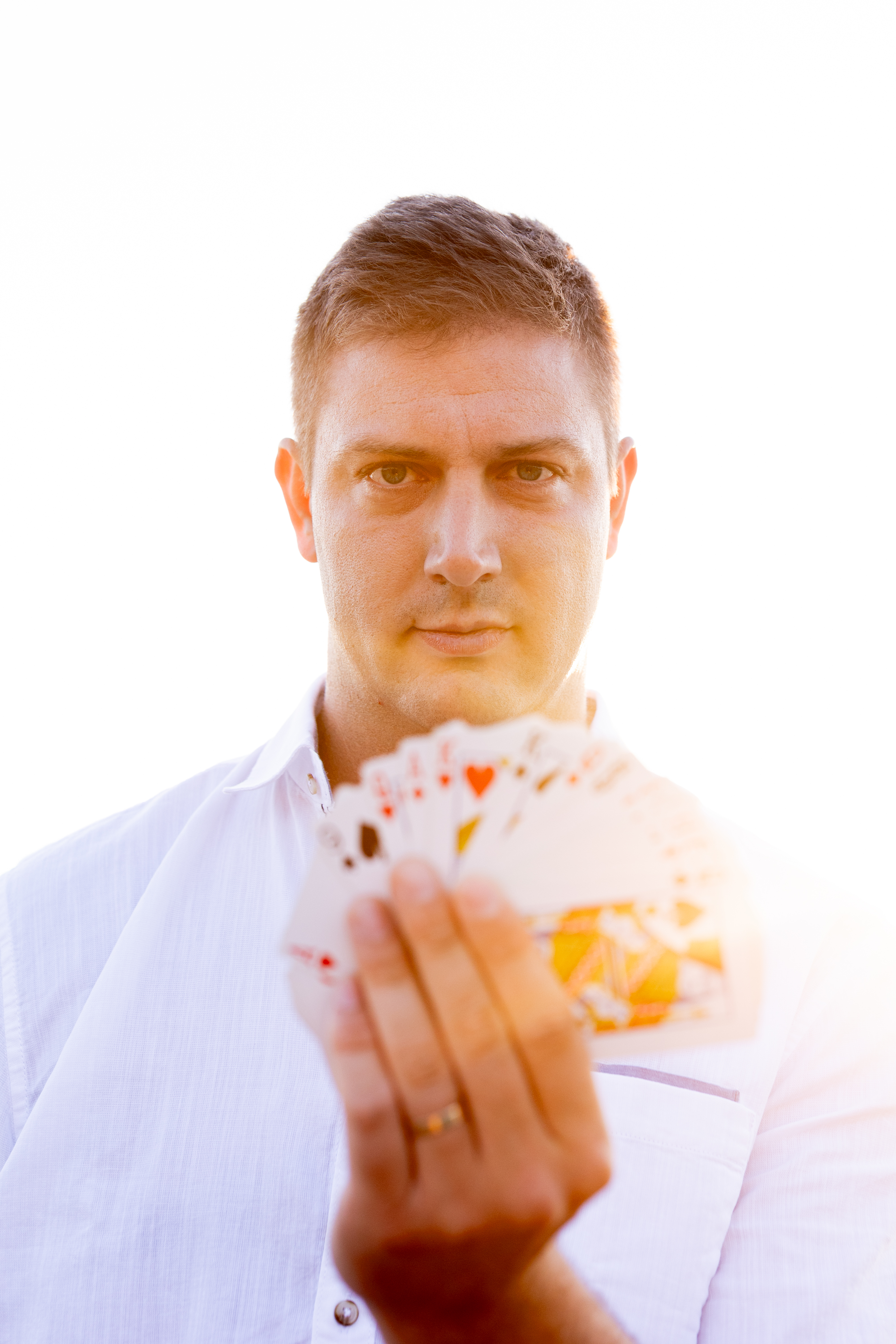 Portrait of a man in a white shirt holding a fanned deck of cards, lit by bright golden daylight, clean and focused.