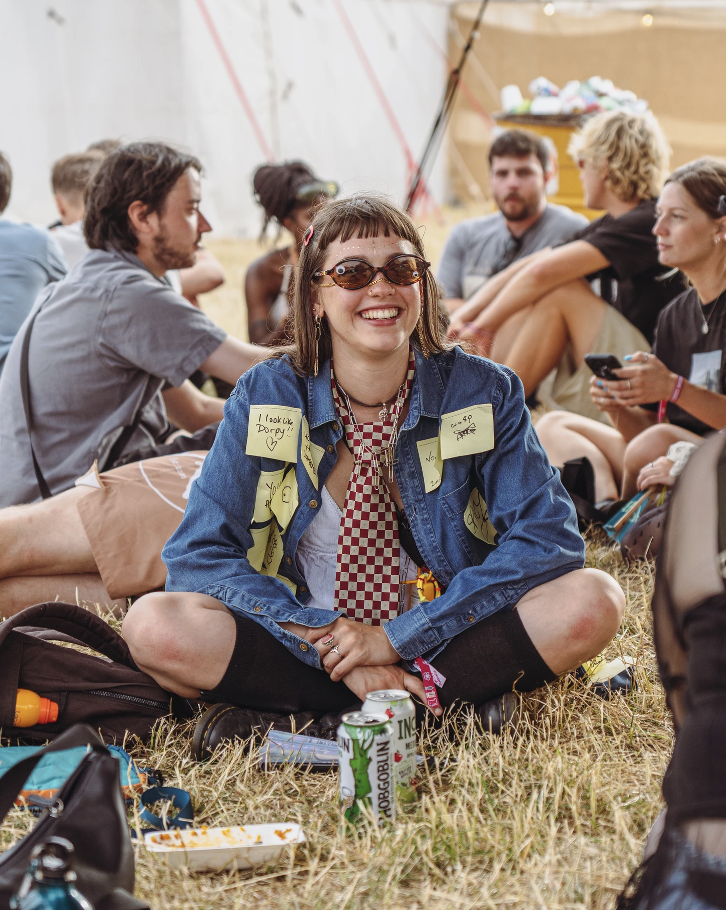 A lively outdoor festival scene features a smiling woman with quirky sunglasses and a denim jacket, surrounded by friends on grass, radiating joy.