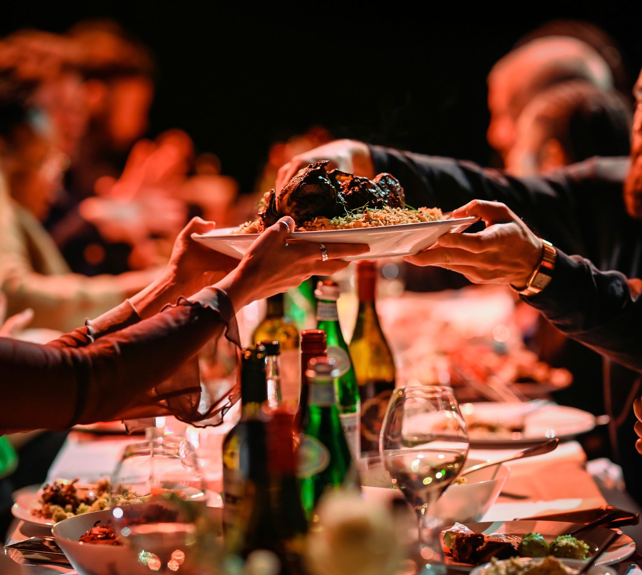In a cozy, warm-lit setting, hands reach for a steaming platter of savory meat atop fragrant rice, flanked by polished glassware and vibrant bottles.
