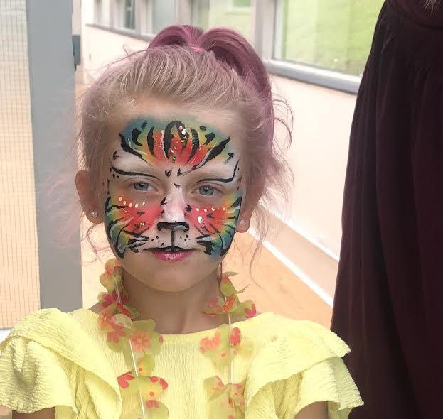 A girl with vibrant tiger face paint and pink hair wears a yellow ruffled top, exuding playful energy in a bright, cheerful space.