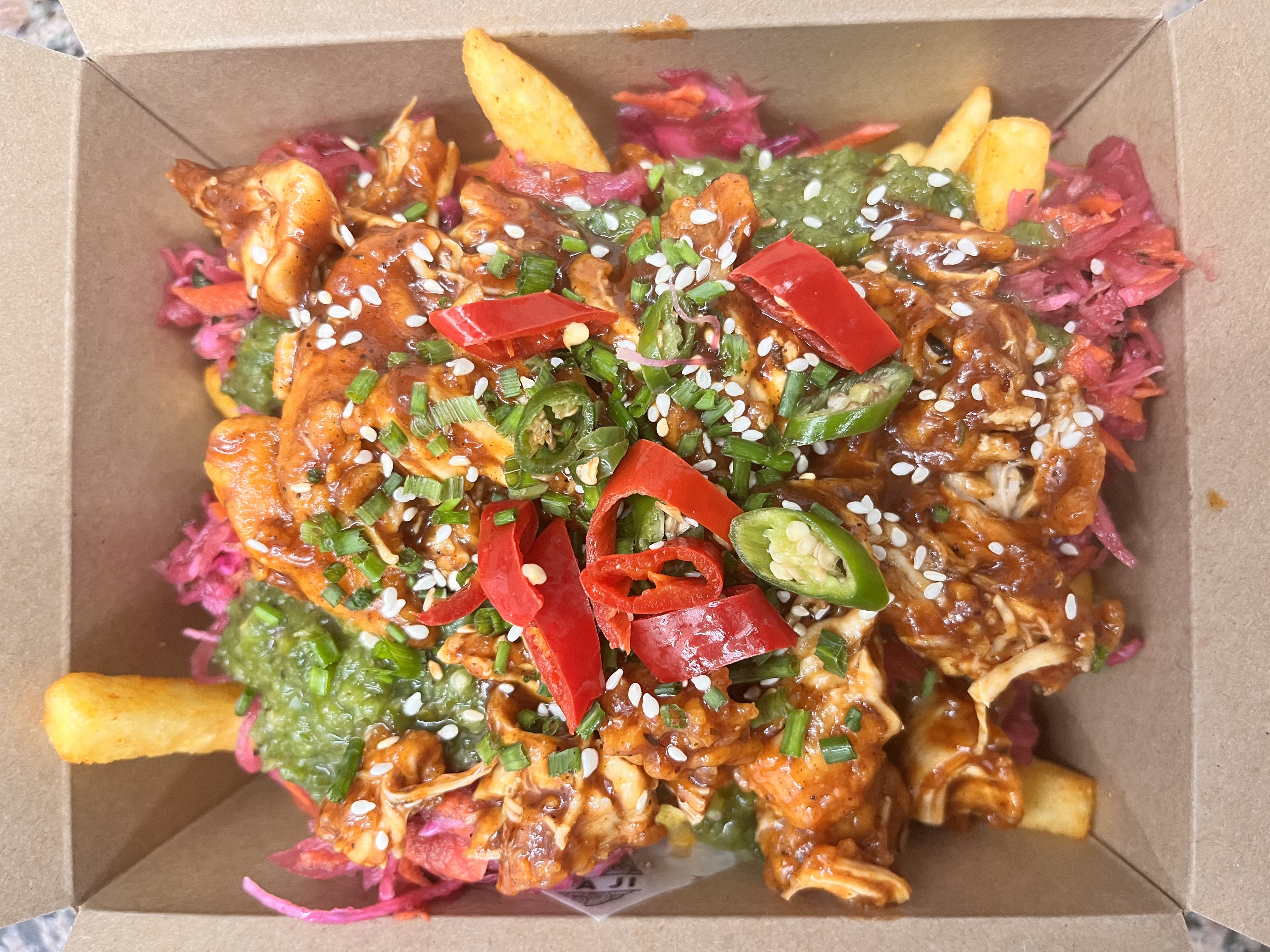 A vibrant mix of crispy fries, spicy chicken, green sauce, and pickled cabbage topped with sesame seeds and sliced chilies in a sleek box.
