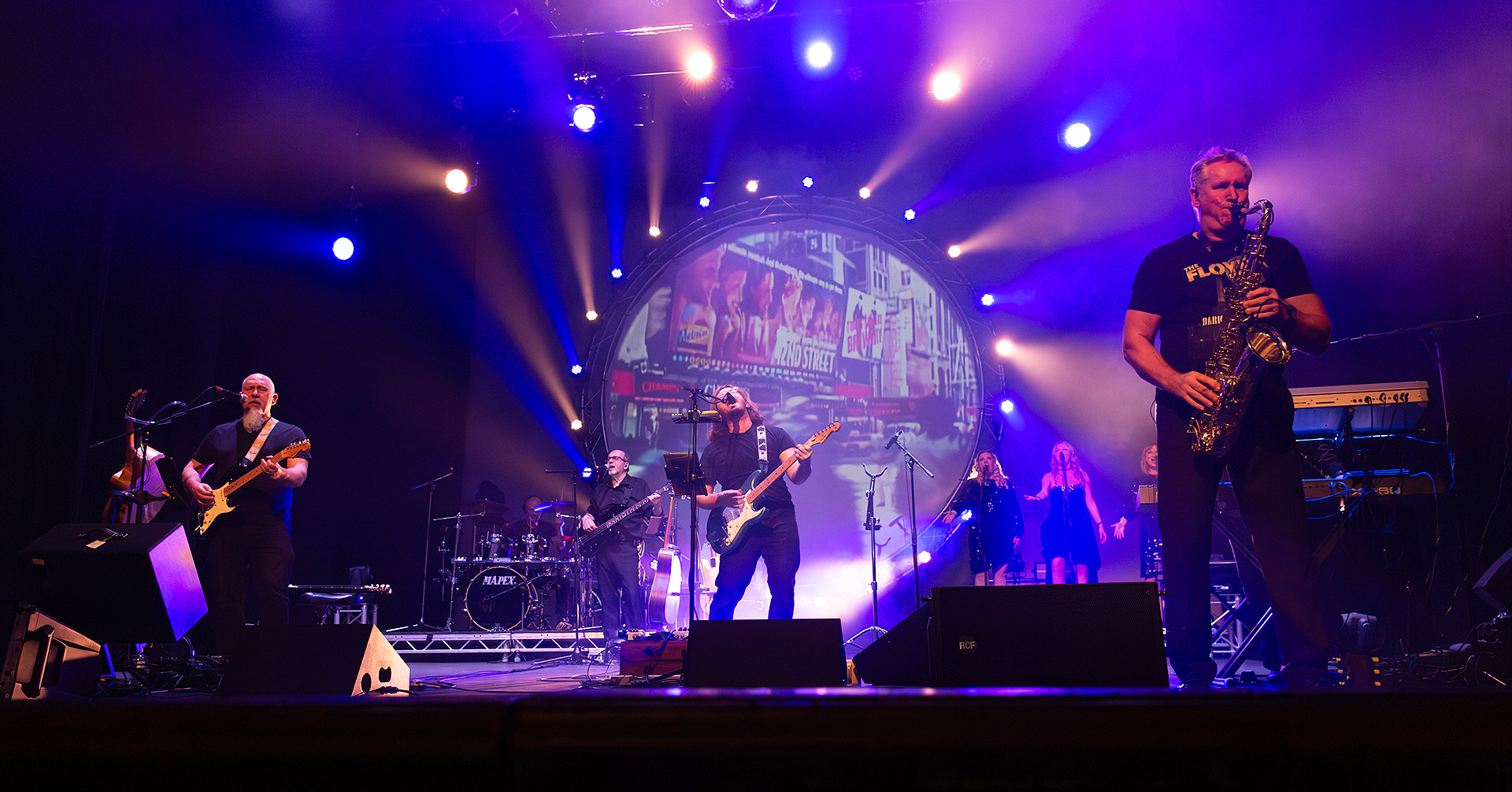 A vibrant band performs on stage, illuminated by colorful lights, with musicians energetically playing instruments, creating a lively atmosphere.