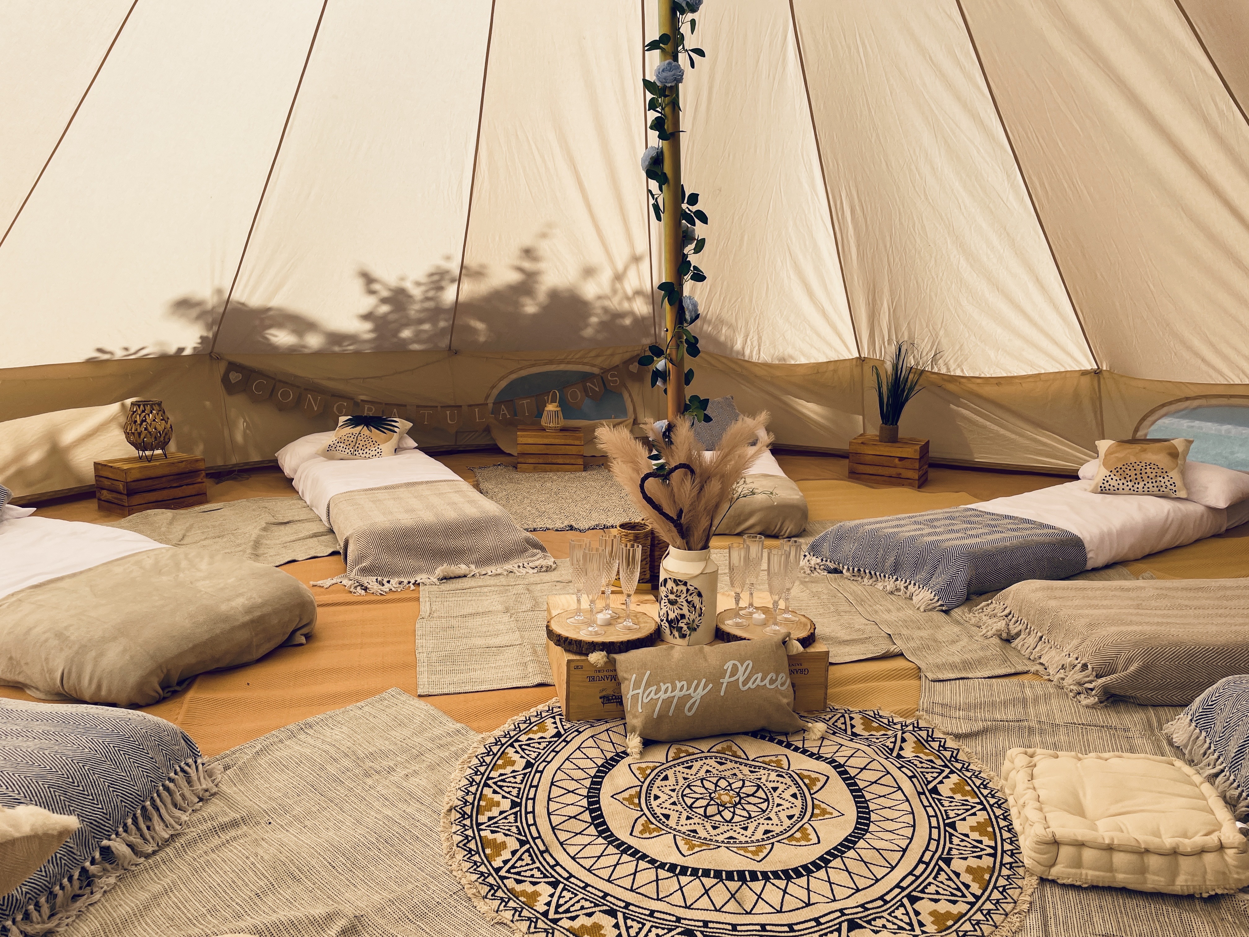 A cozy tent setting features plush, light beige cushions arranged on woven rugs, with gentle warm lighting and rustic wooden accents.