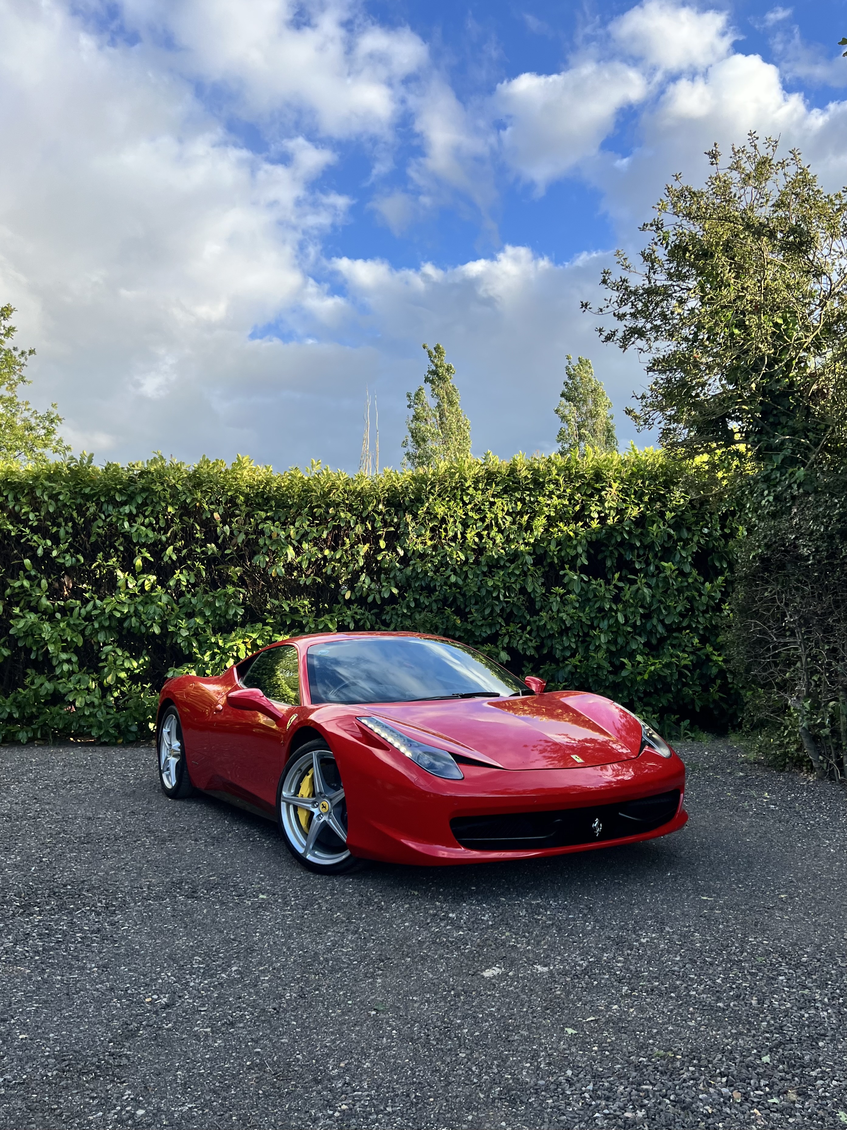 A glossy red Ferrari coupe sits on a gravel drive before manicured hedges, under bright daylight and a vivid blue, cloud-filled sky.