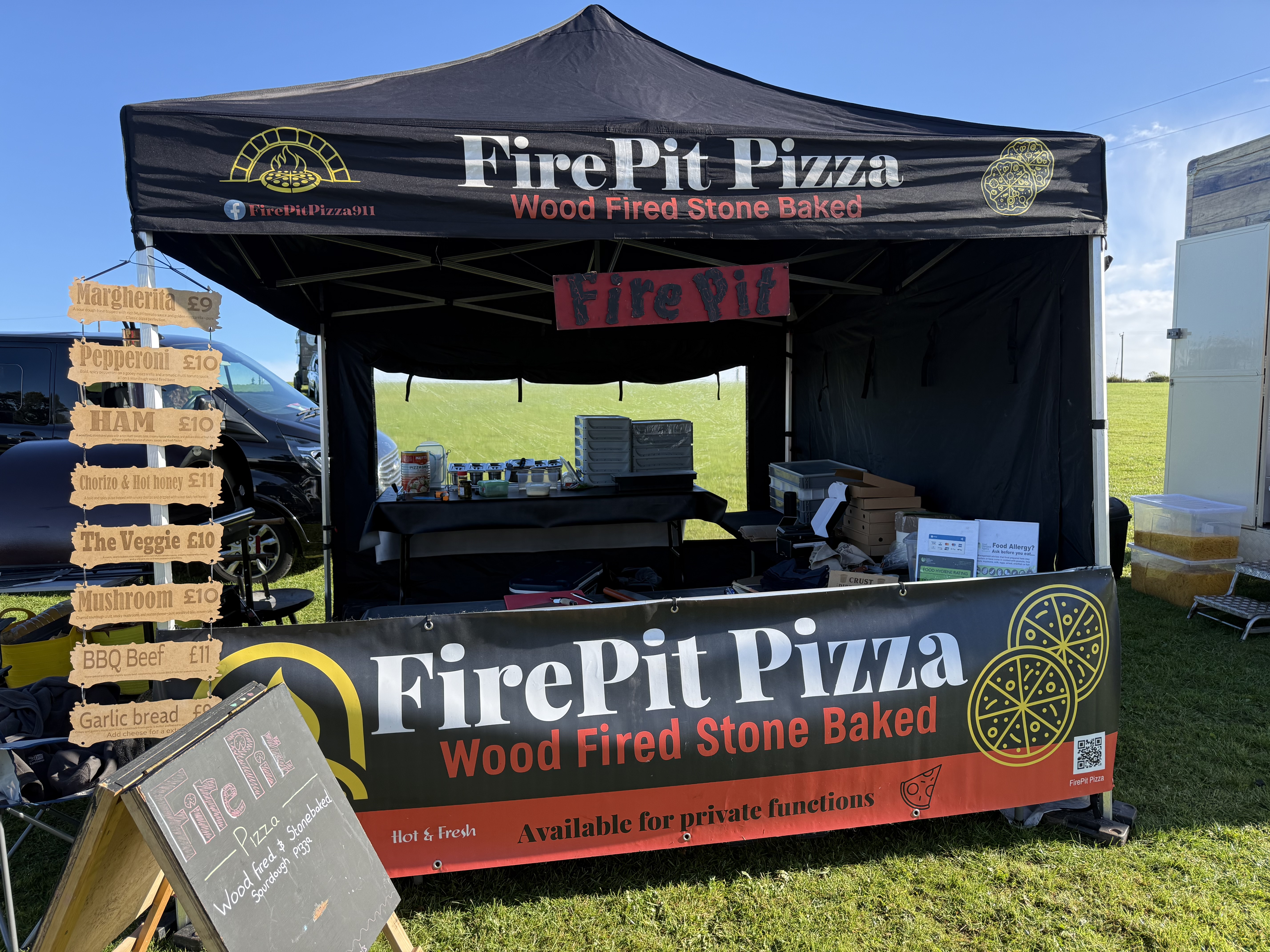 The vibrant FirePit Pizza stall features a black canopy, wooden menu signs showcasing pizza options, and a clean, inviting atmosphere, perfect for outdoor catering.