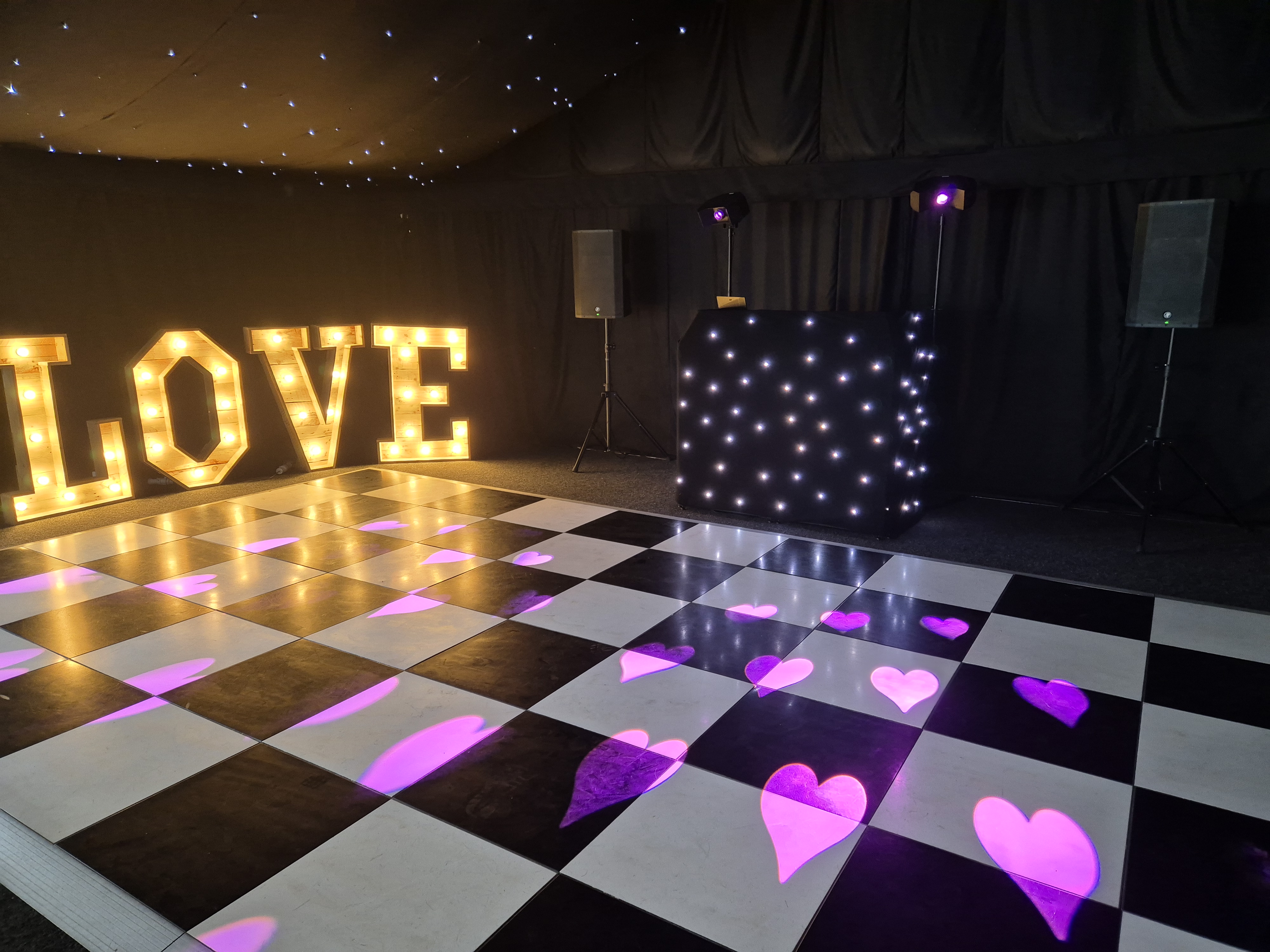 Starry black-walled wedding dance floor with checkered tiles, glowing LOVE marquee, LED DJ booth, and pink heart lights under warm spotlight.