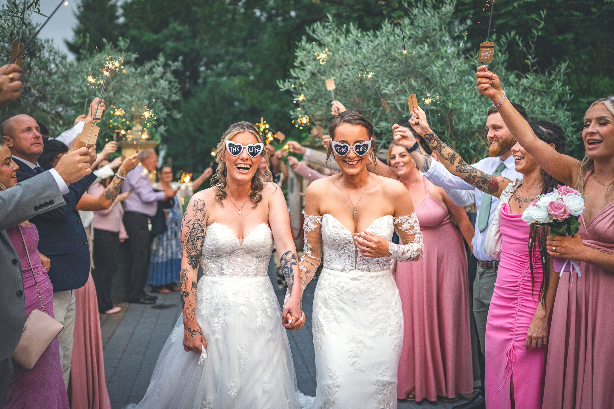 Two brides in lace gowns and “The Wife” sunglasses walk through a sparkler tunnel, with cheering guests, pink dresses, and warm evening wedding vibes.