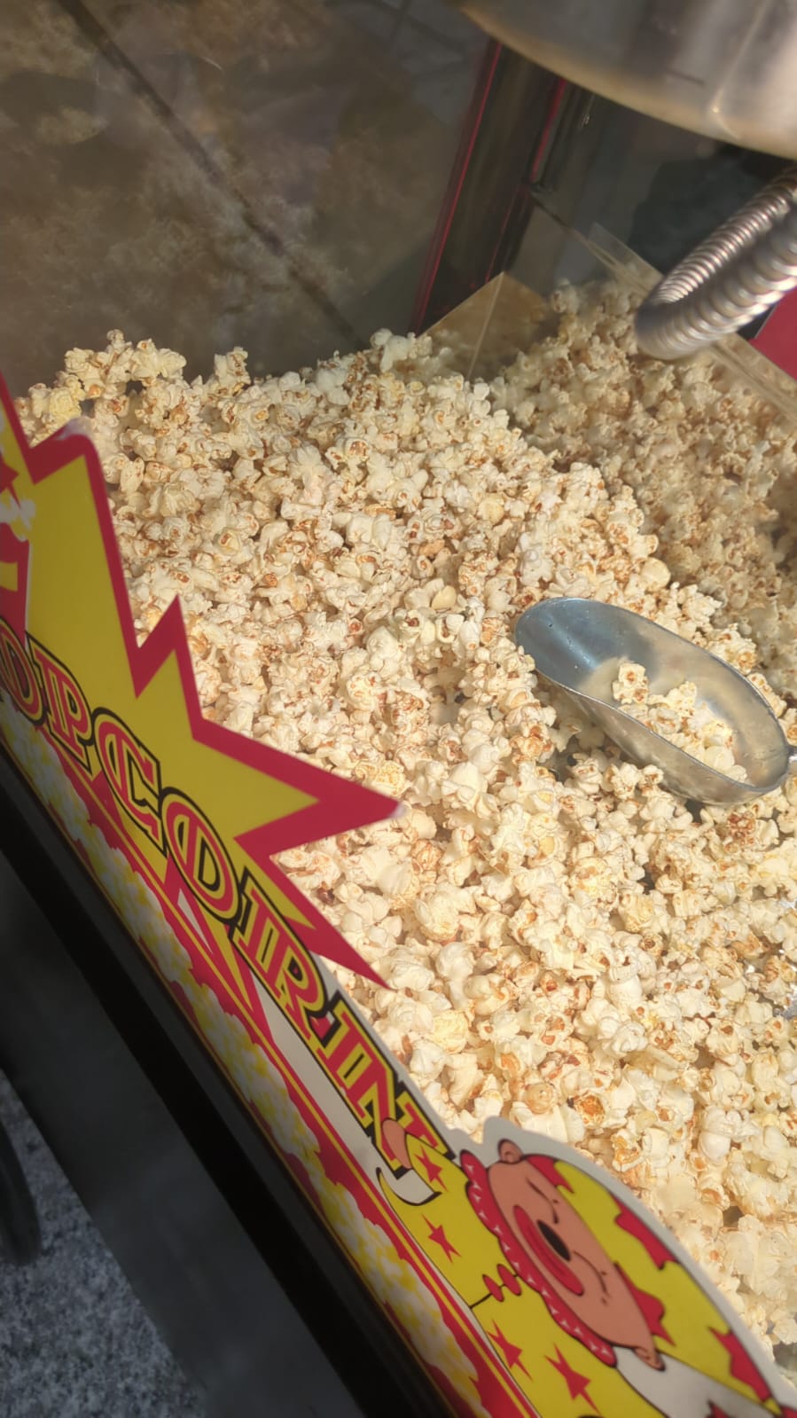 Bright popcorn catering display in a glass-topped cart, with buttery kernels piled high, silver scoop, and bold yellow-red signage under warm event lighting.