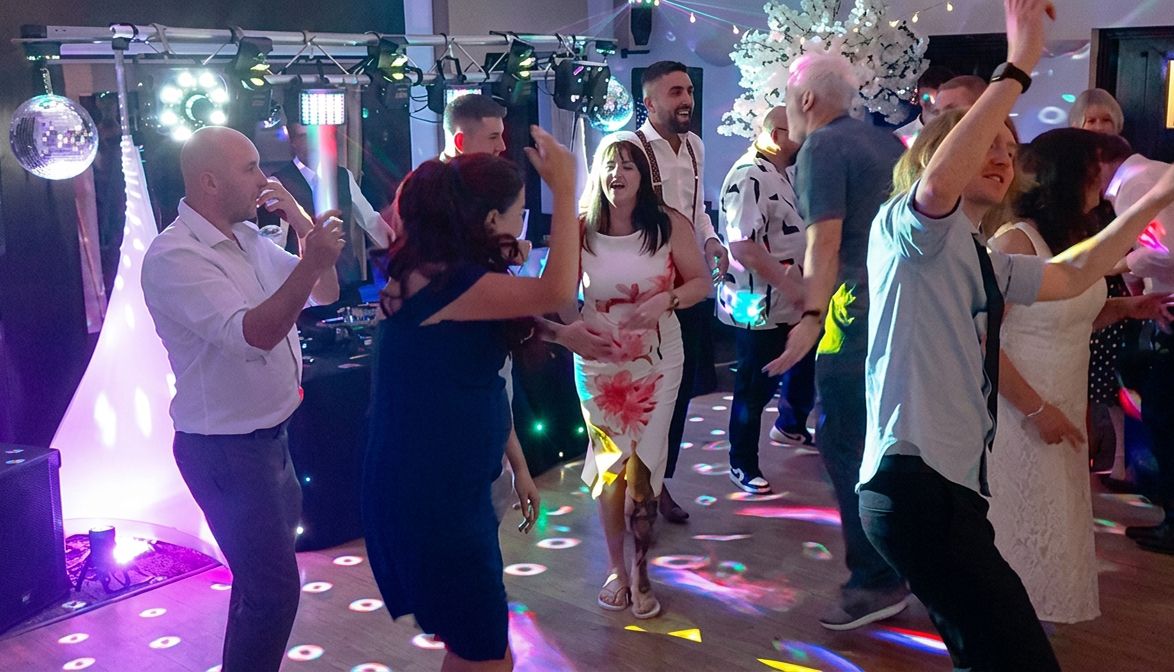 Energetic wedding reception dance floor with DJ lights, disco balls, and guests in semi-circle, vibrant party atmosphere.