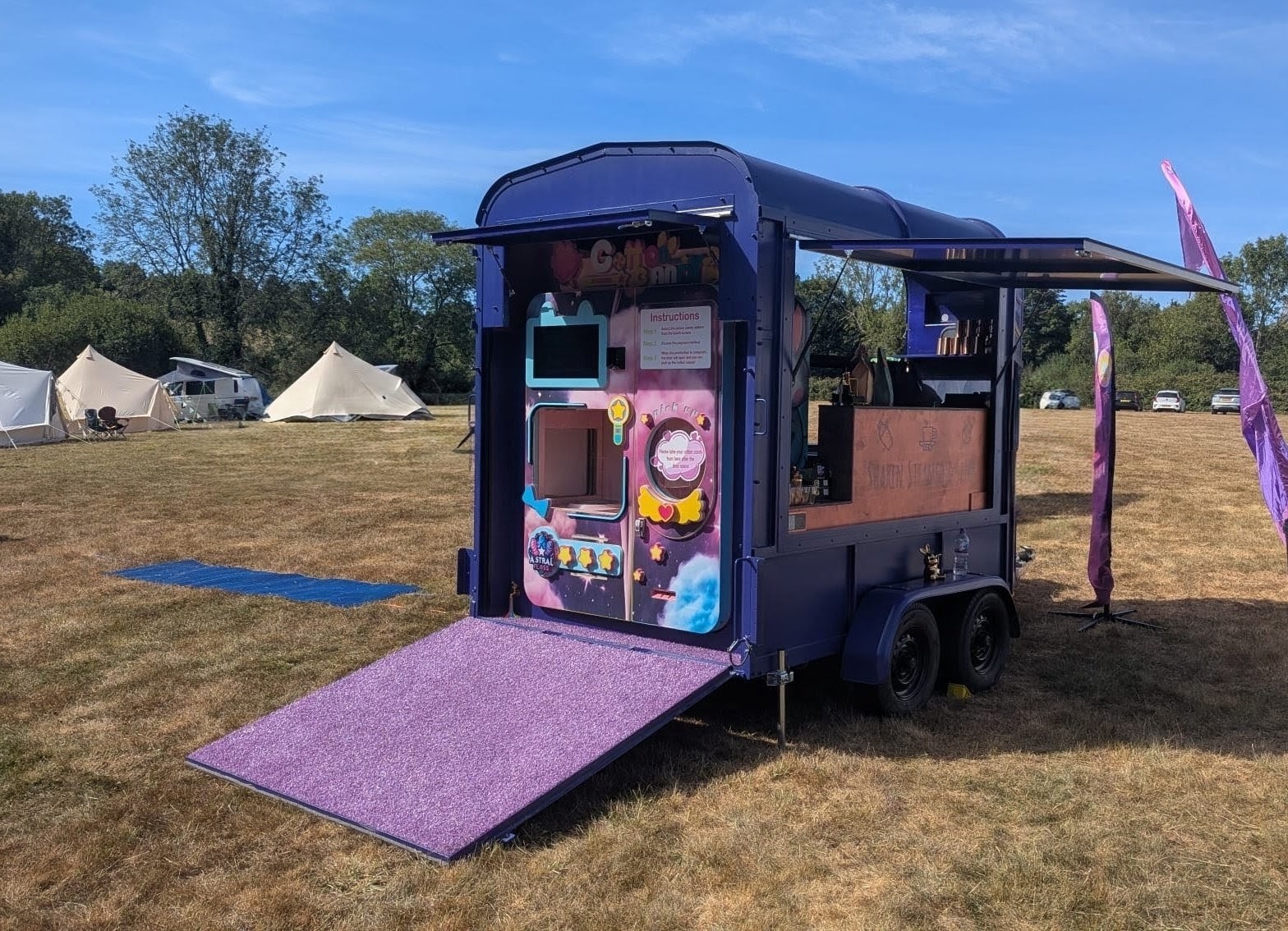 Purple catering trailer with glitter ramp and awnings on a sunny field, clean festival setup beside tents and flags, polished service.