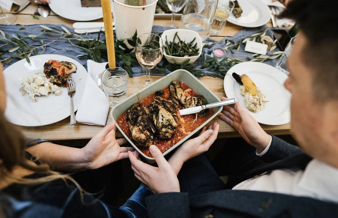 A rustic wooden table displays roasted chicken with crispy skin in tomato sauce, vibrant green beans, and elegant glassware under warm lighting.