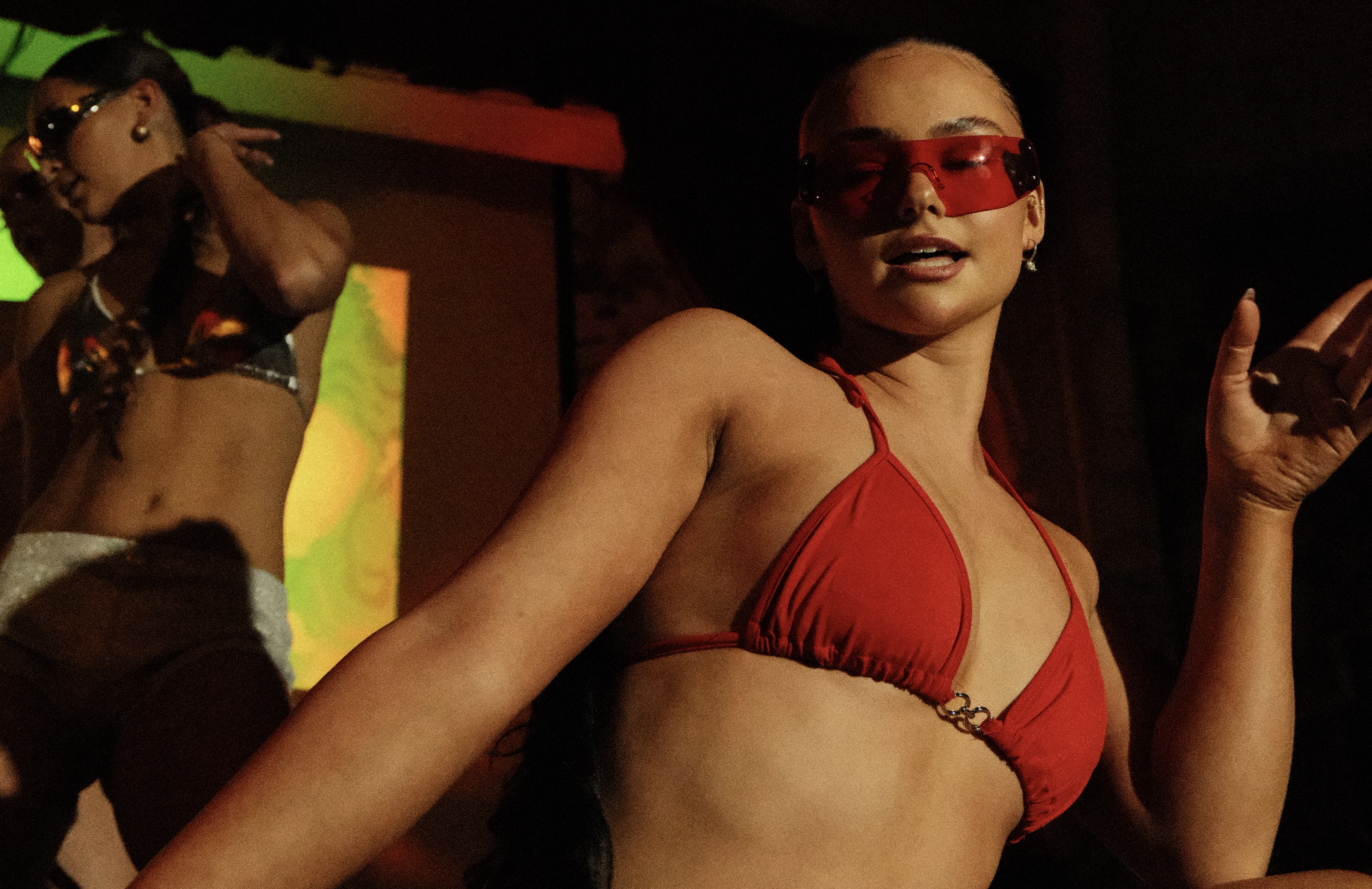 A vibrant nightlife scene features two women dancing energetically, adorned in bold swimwear, with dynamic lighting enhancing the electric atmosphere.