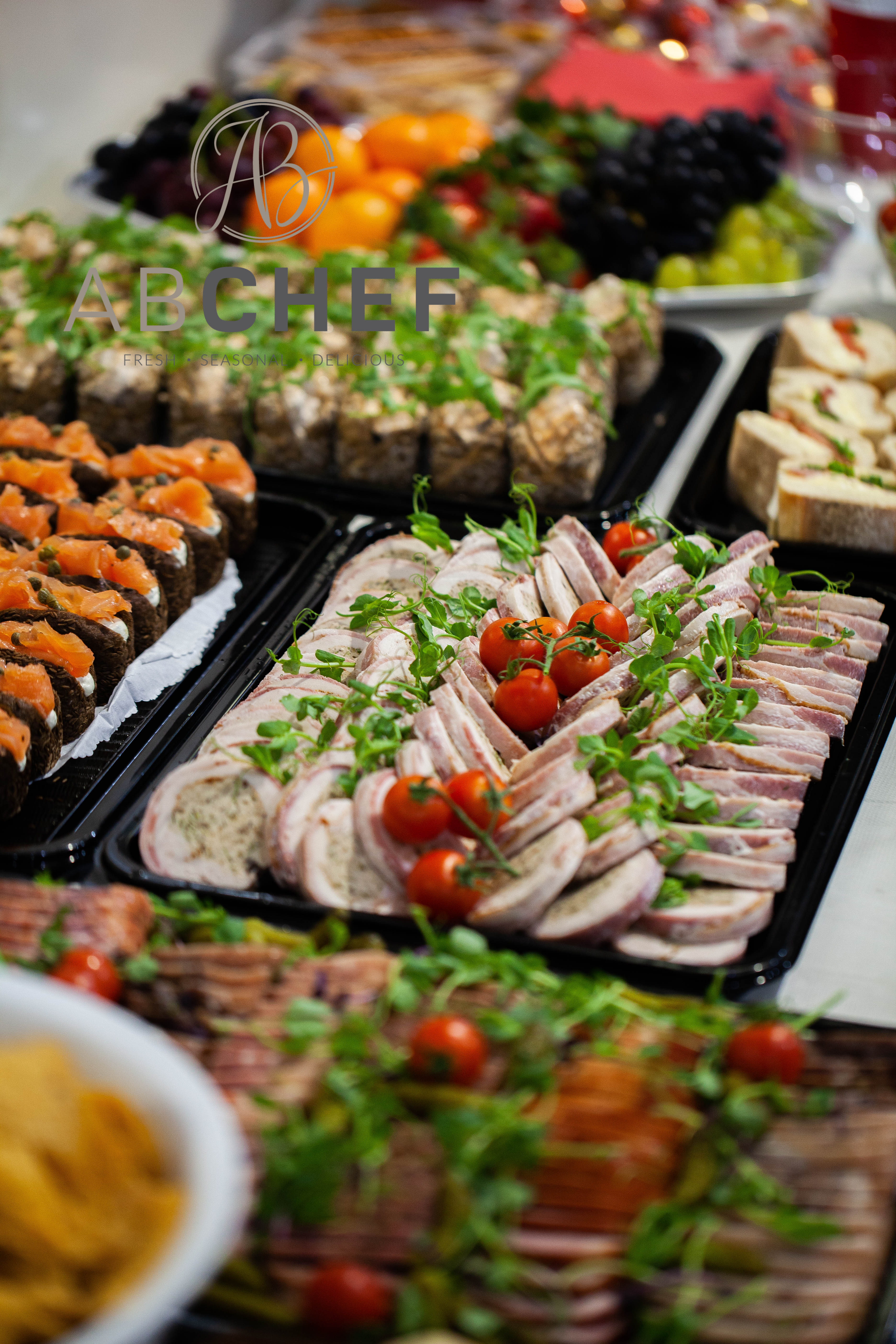 An elegant catering display features neatly arranged smoked salmon bites, delicately sliced meats, vibrant cherry tomatoes, and fresh greens, set on glossy black trays, creating a festive, appetizing atmosphere.