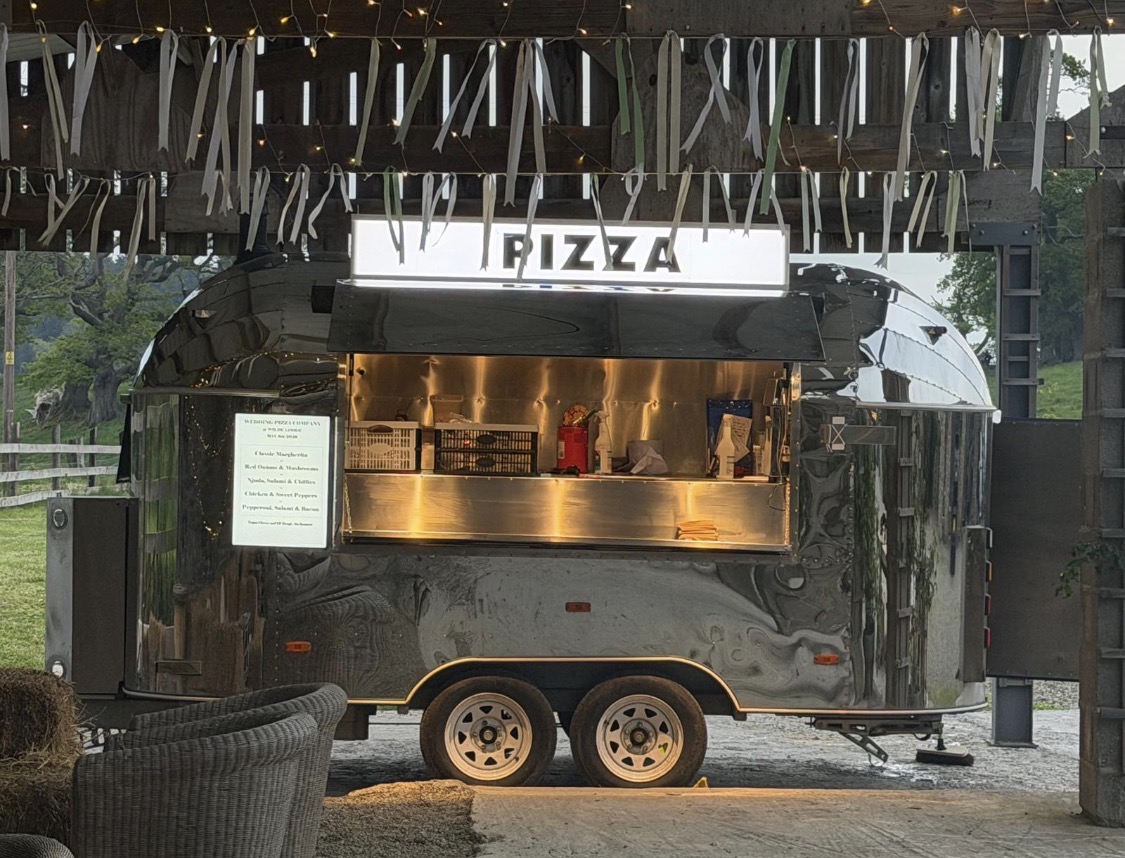 A sleek silver pizza van glows under warm evening lights, showcasing fresh ingredients while a staff member prepares meals in an inviting atmosphere.