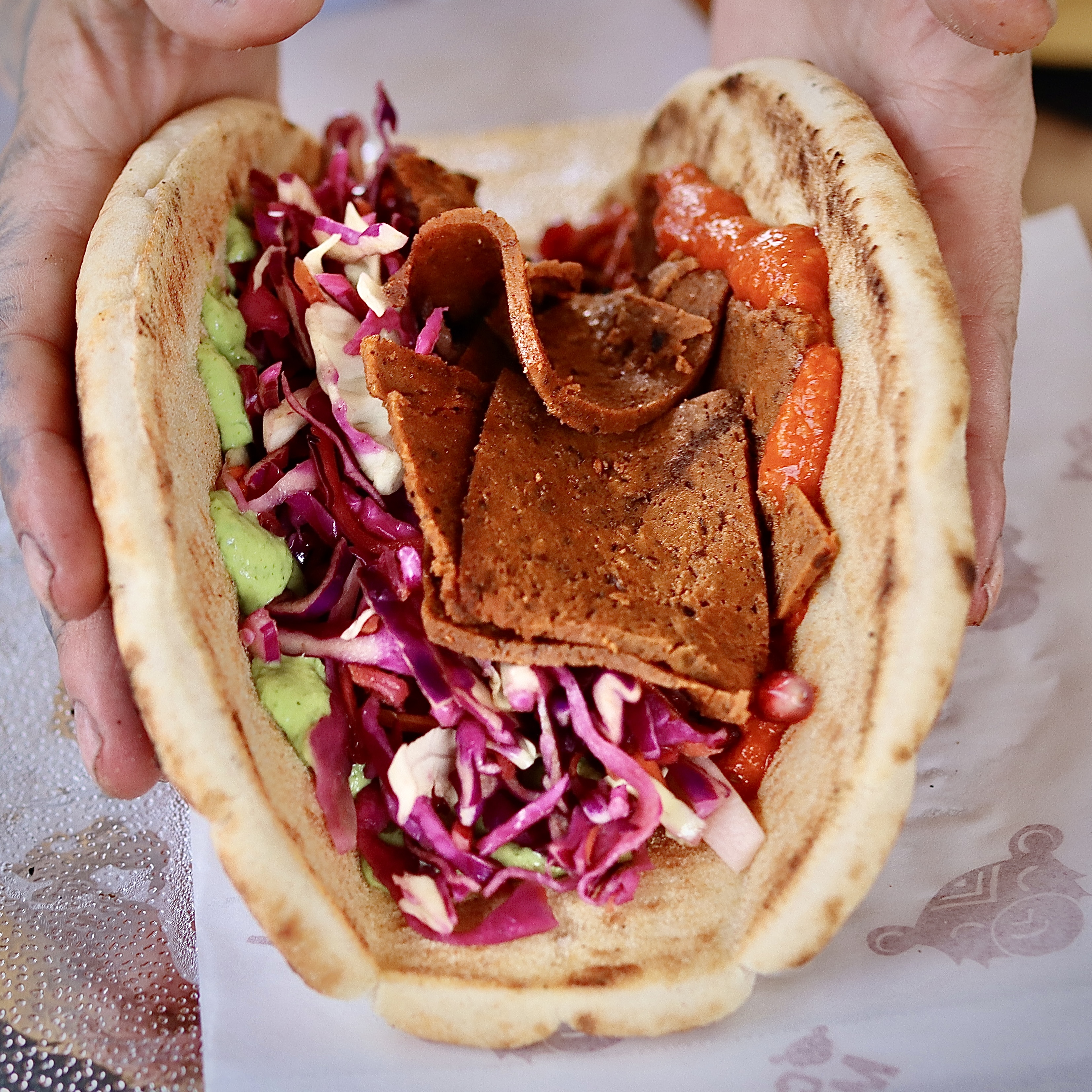 A hand holds a warm pita stuffed with vibrant red cabbage, green sauce, and spiced meat, showcasing fresh, artful street food presentation.