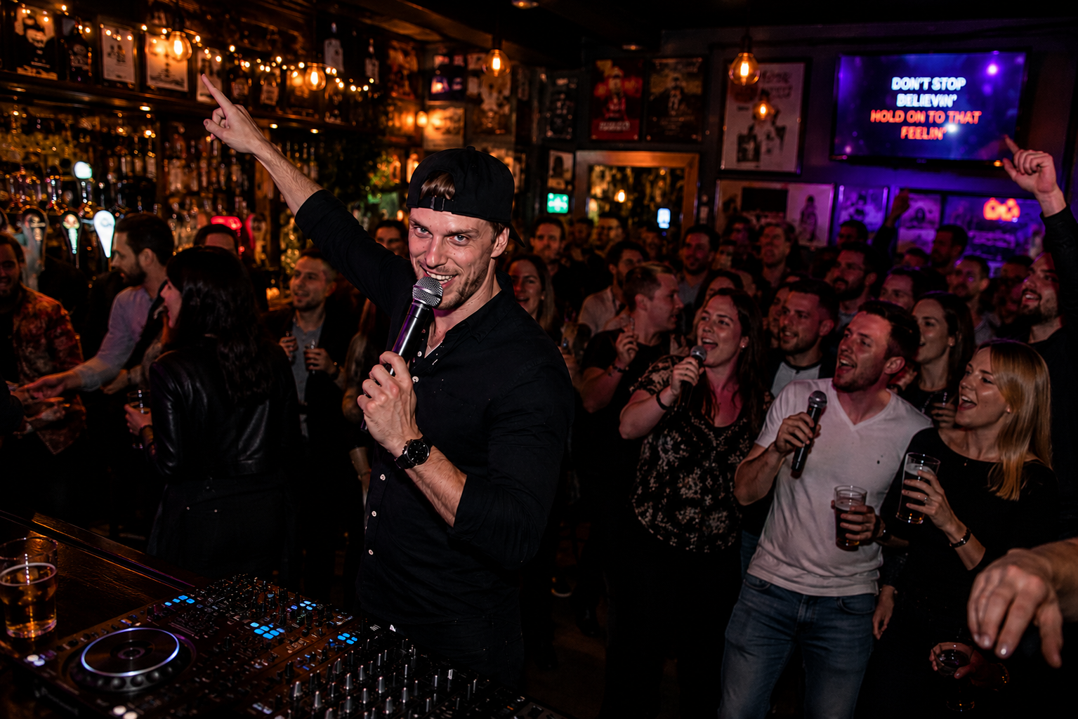 Energetic karaoke night in a packed neon-lit bar, DJ deck in foreground, singers with microphones and drinks, lively catering atmosphere.