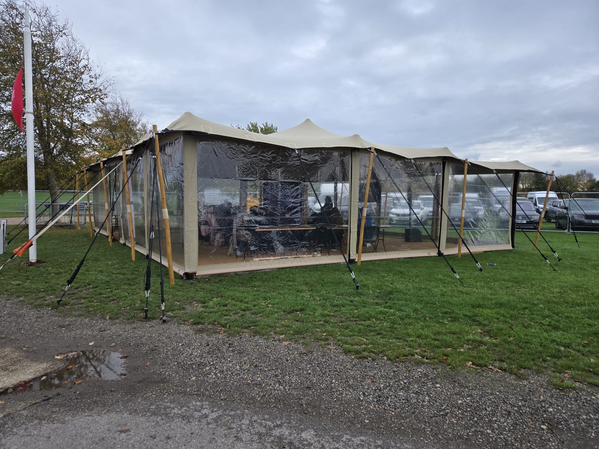 A spacious tent with transparent panels and wooden supports sits on grassy ground, featuring simple tables and chairs for a cozy, casual setting.