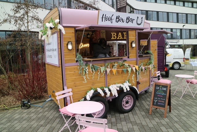 A charming, vintage-inspired bar trailer with pink accents and wooden panels offers drinks, inviting patrons to gather at round tables surrounded by greenery.