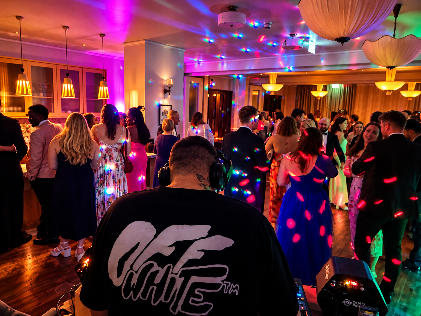 Crowded wedding reception with DJ in black tee, colorful disco lights, guests in formalwear, warm pendant lighting, polished dance floor, lively catering vibe.