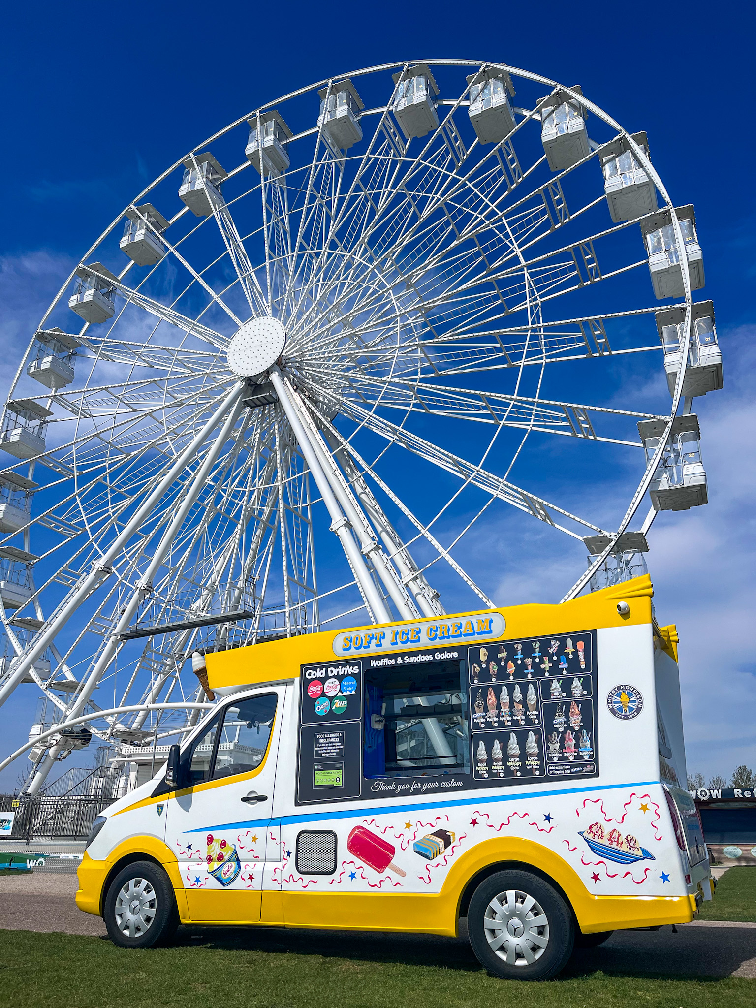 A vibrant ice cream van in front of a large, white Ferris wheel against a clear blue sky, inviting guests to enjoy sweet treats outdoors.