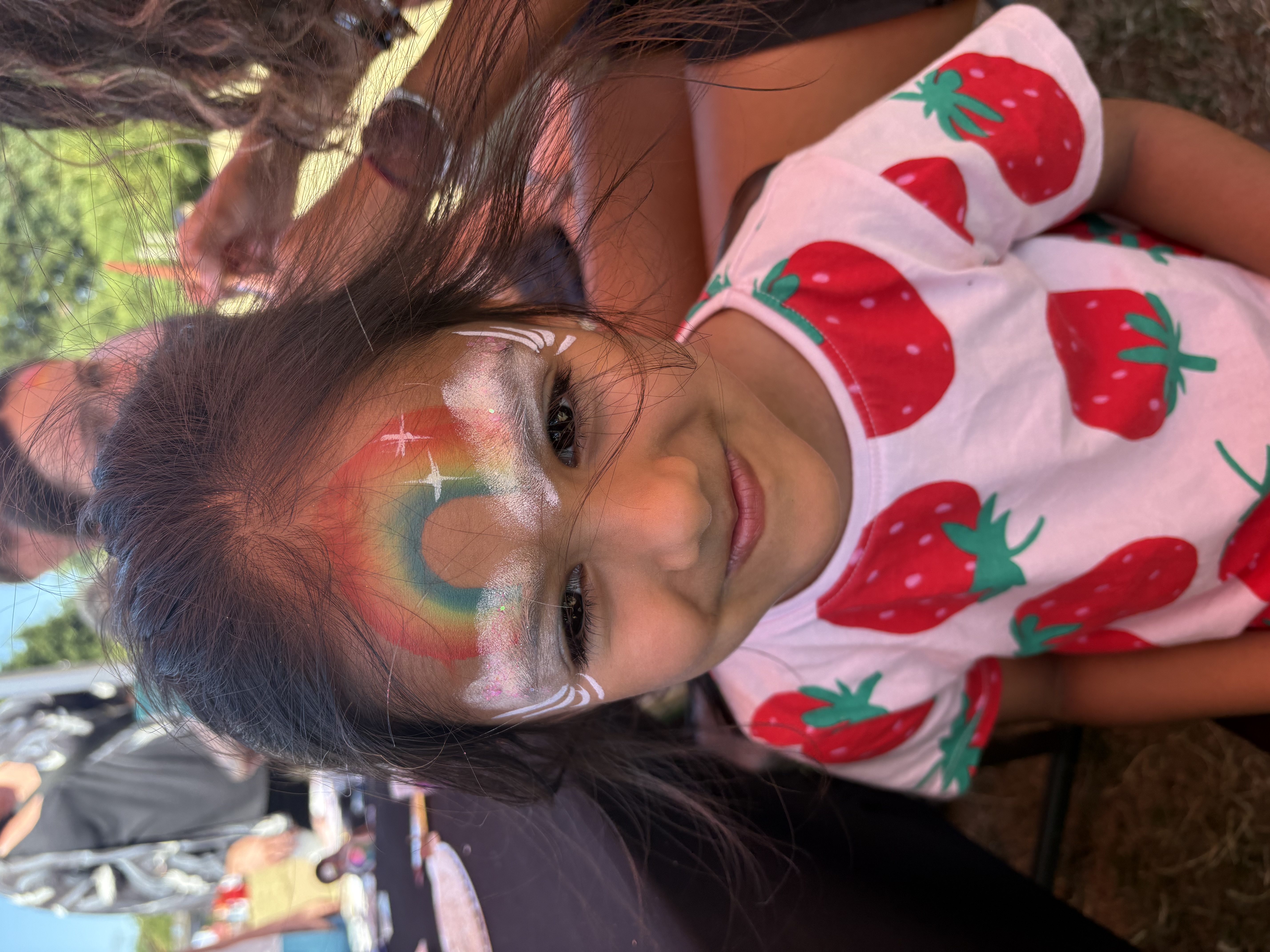 A joyful child with a rainbow face painting smiles, wearing a strawberry-patterned shirt, amidst a vibrant outdoor festival atmosphere.