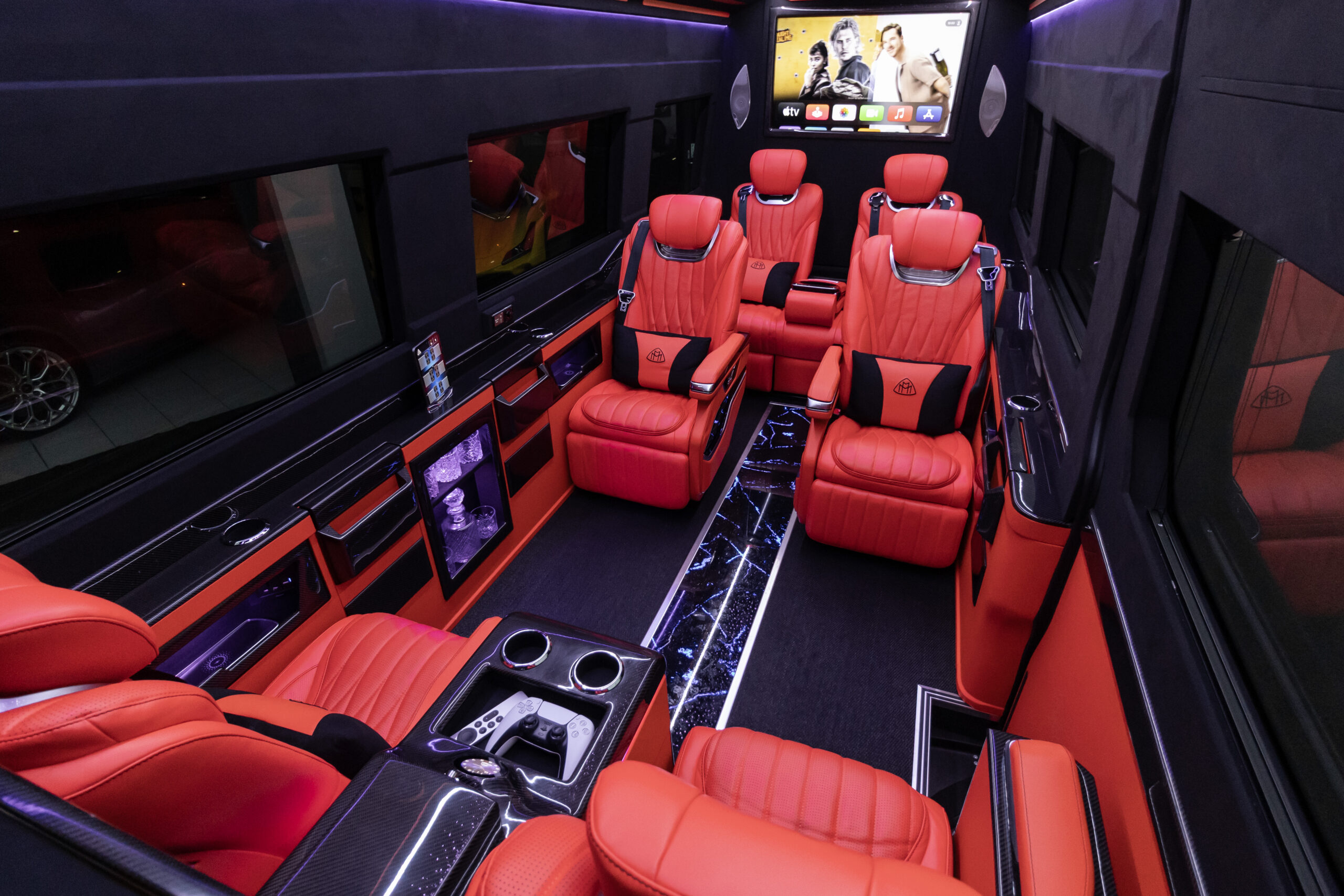 Luxury limo bus interior with red leather captain seats, black accents, LED strip lighting, glossy consoles, and polished AV screens.