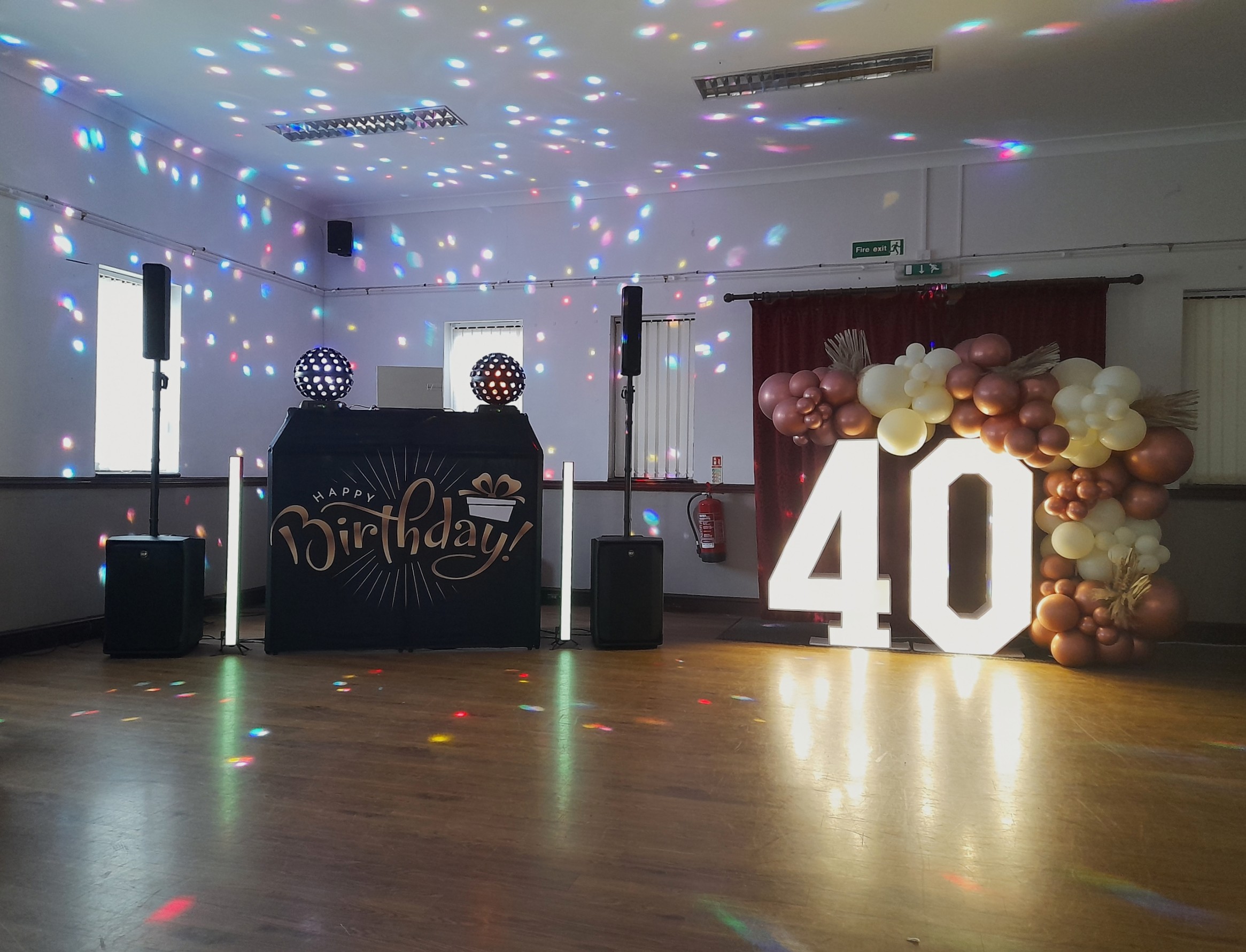 Birthday DJ setup with disco lights, black speakers, illuminated 40 sign, rose-gold balloon arch, and polished wood floor.