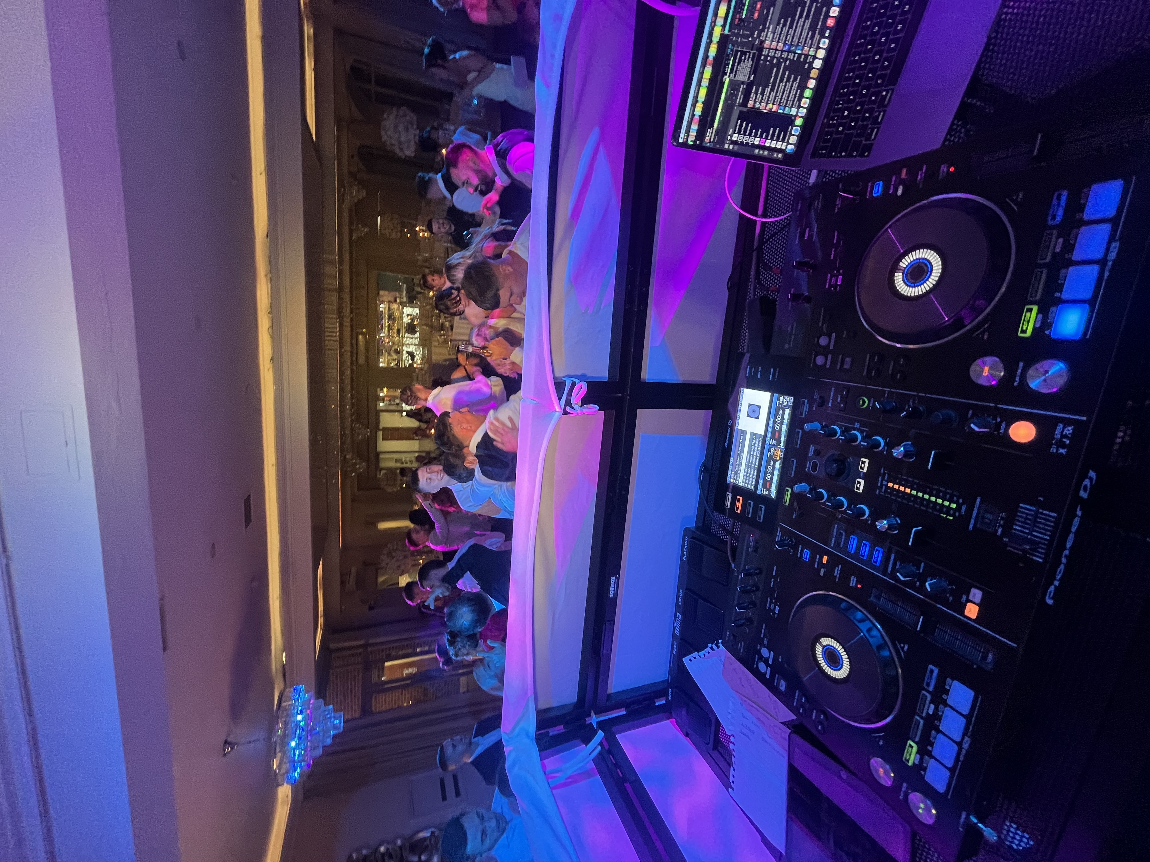 Vibrant wedding DJ booth with Pioneer decks and laptop foreground, facing a lively crowded dance floor under purple-blue uplighting.