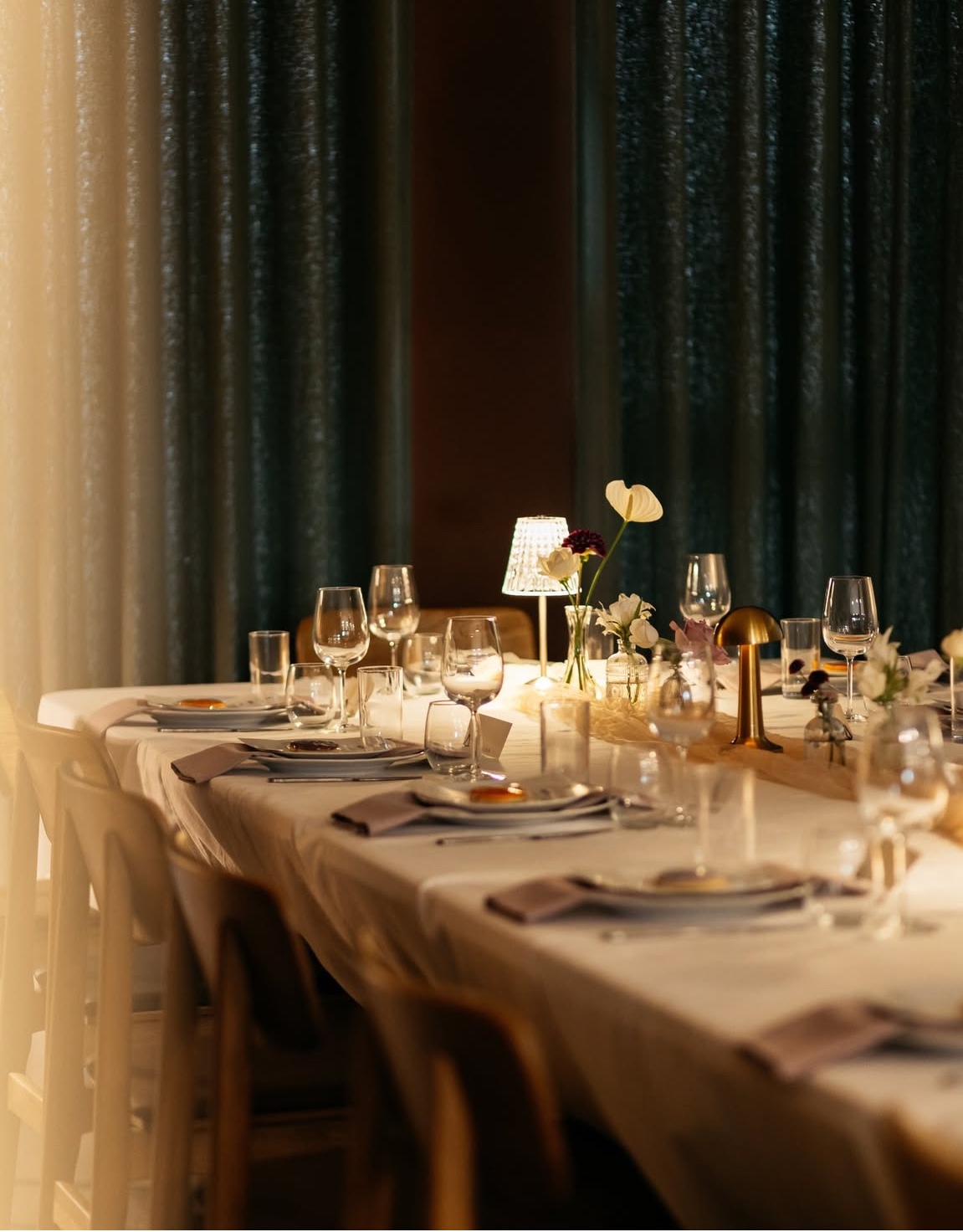 A long, elegantly set table features polished glassware and soft linen napkins under warm lighting, adorned with delicate flowers, exuding a refined atmosphere.