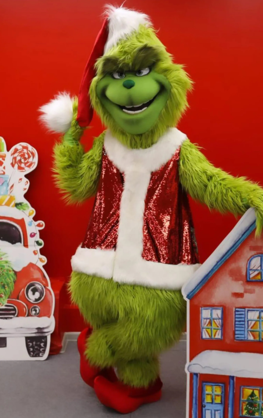 A vibrant red backdrop sets a playful scene with the Grinch wearing a sparkly vest, waving cheerfully next to a whimsical house cutout.