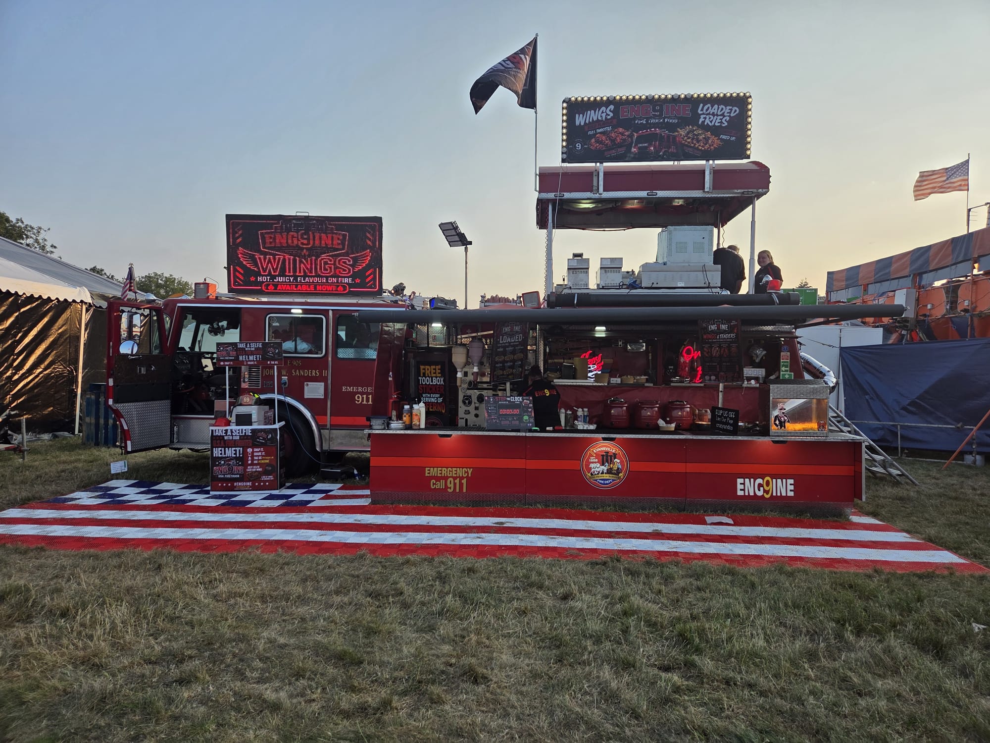 Red fire-engine food truck serving wings and loaded fries, arranged in a bold festival setup with neon signs and warm sunset light.