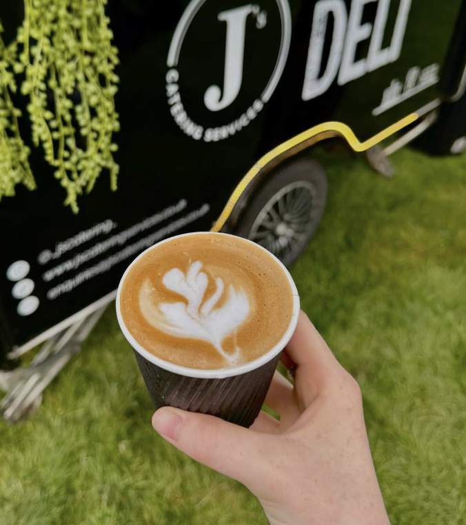 Handheld latte with heart latte art in a black cup, beside a stylish mobile coffee catering van on bright green grass.
